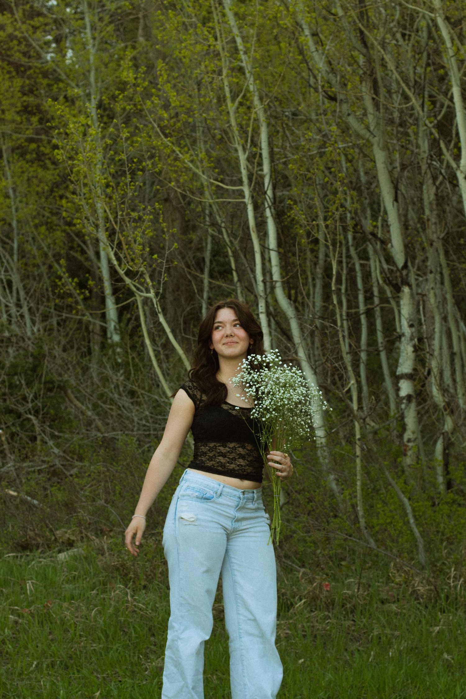 Person in light blue jeans and black top holds delicate white flowers while standing in a forest of birch trees.