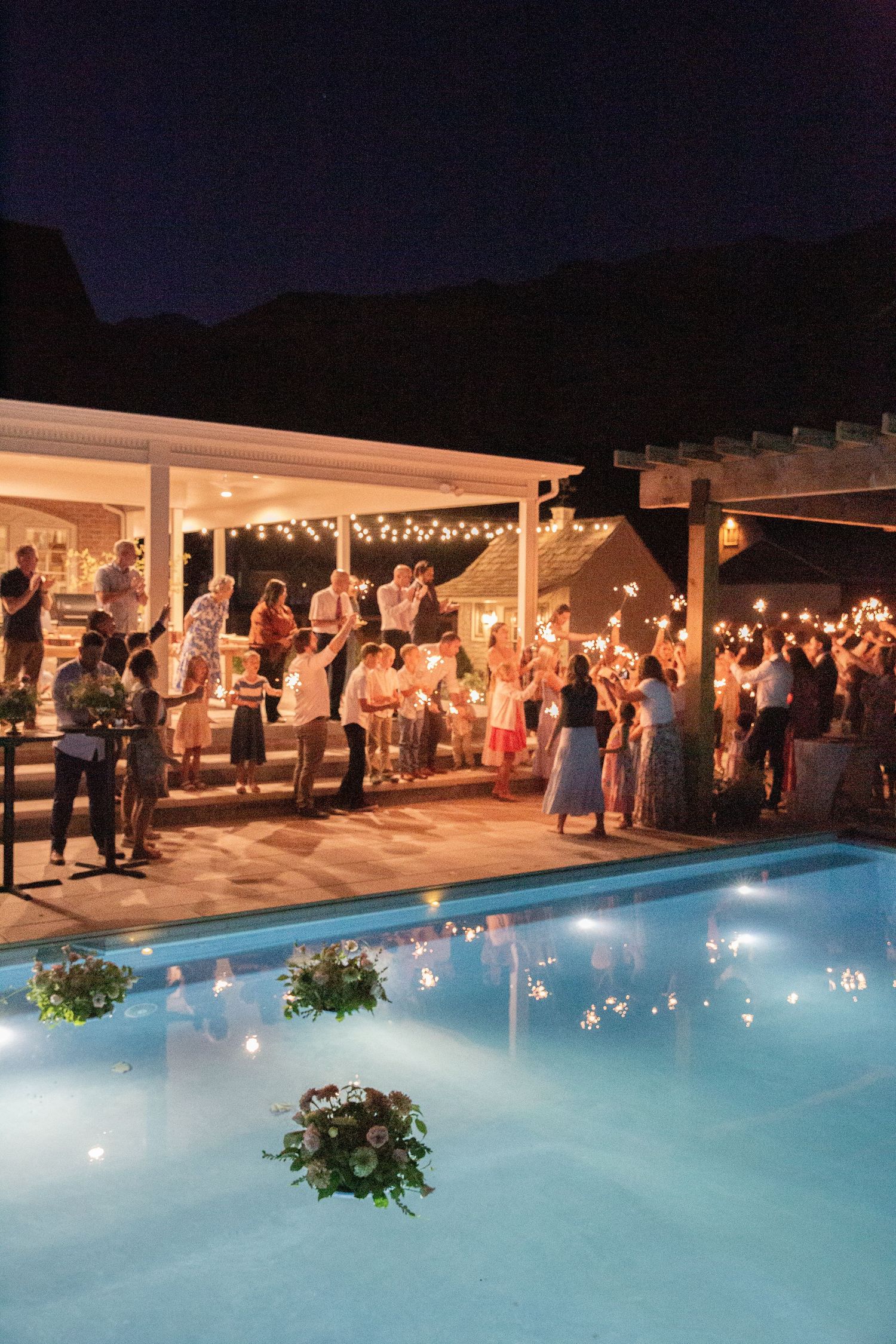 Evening celebration by an illuminated pool with guests mingling under string lights and covered pavilion.