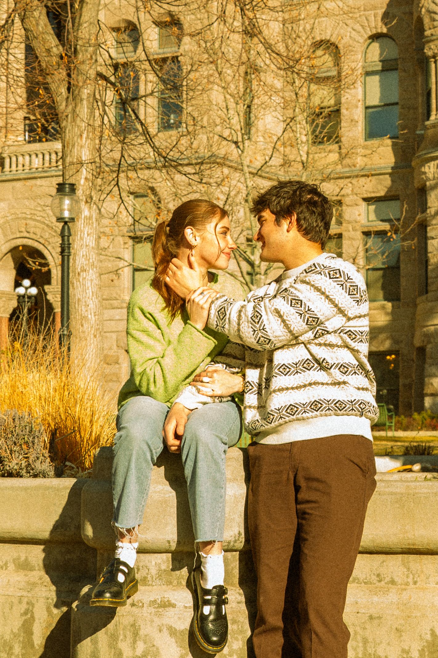 A couple in casual clothing shares a romantic moment against a stone building backdrop in warm autumn sunlight.