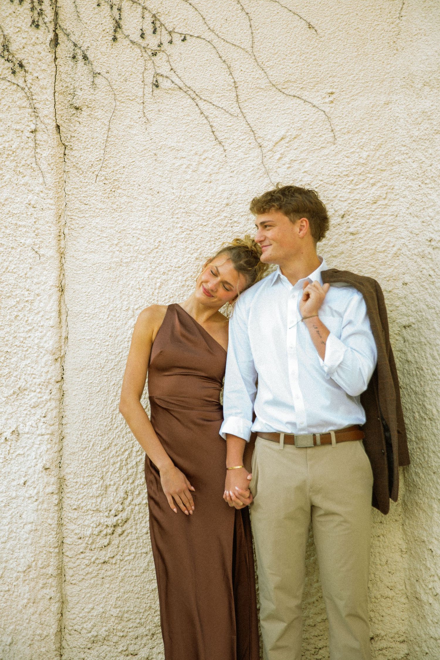 A couple in casual attire stands against a textured white wall, holding hands and sharing an intimate moment together.
