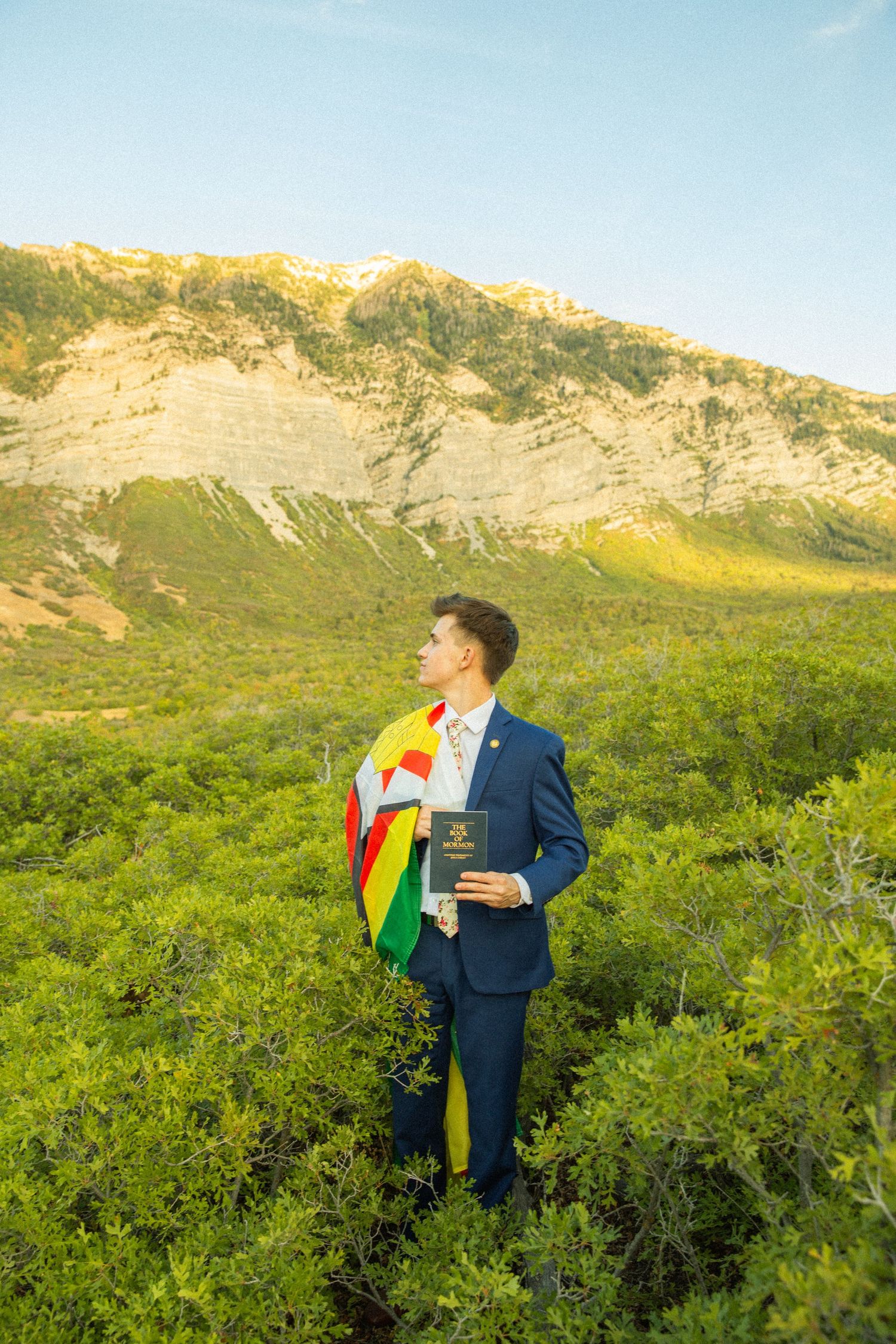 Person in navy suit with rainbow pride flag stands against scenic mountain landscape and green foliage backdrop.