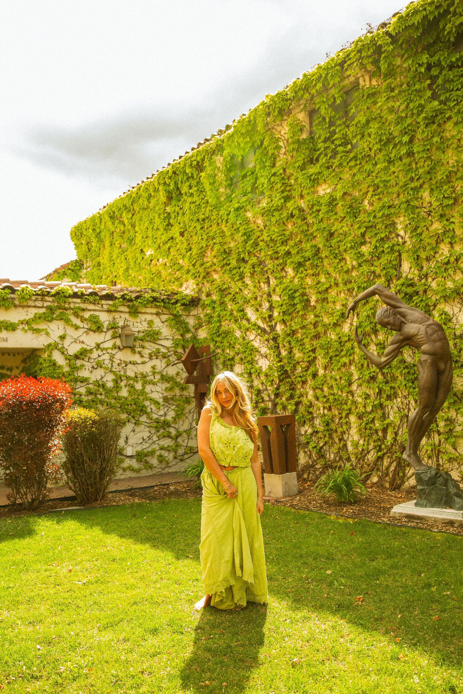 Woman in elegant yellow-green maxi dress posing in garden courtyard with ivy-covered stone wall and bronze sculptures.
