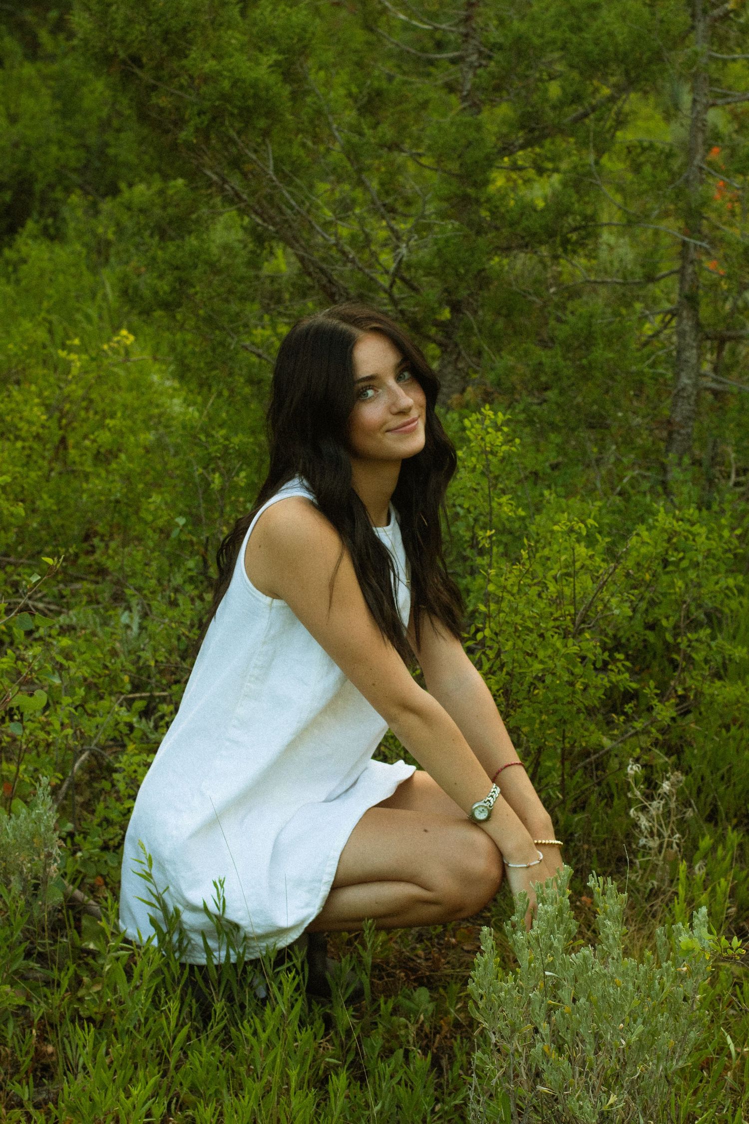 A candid portrait in nature showing someone seated in white clothing among green foliage.