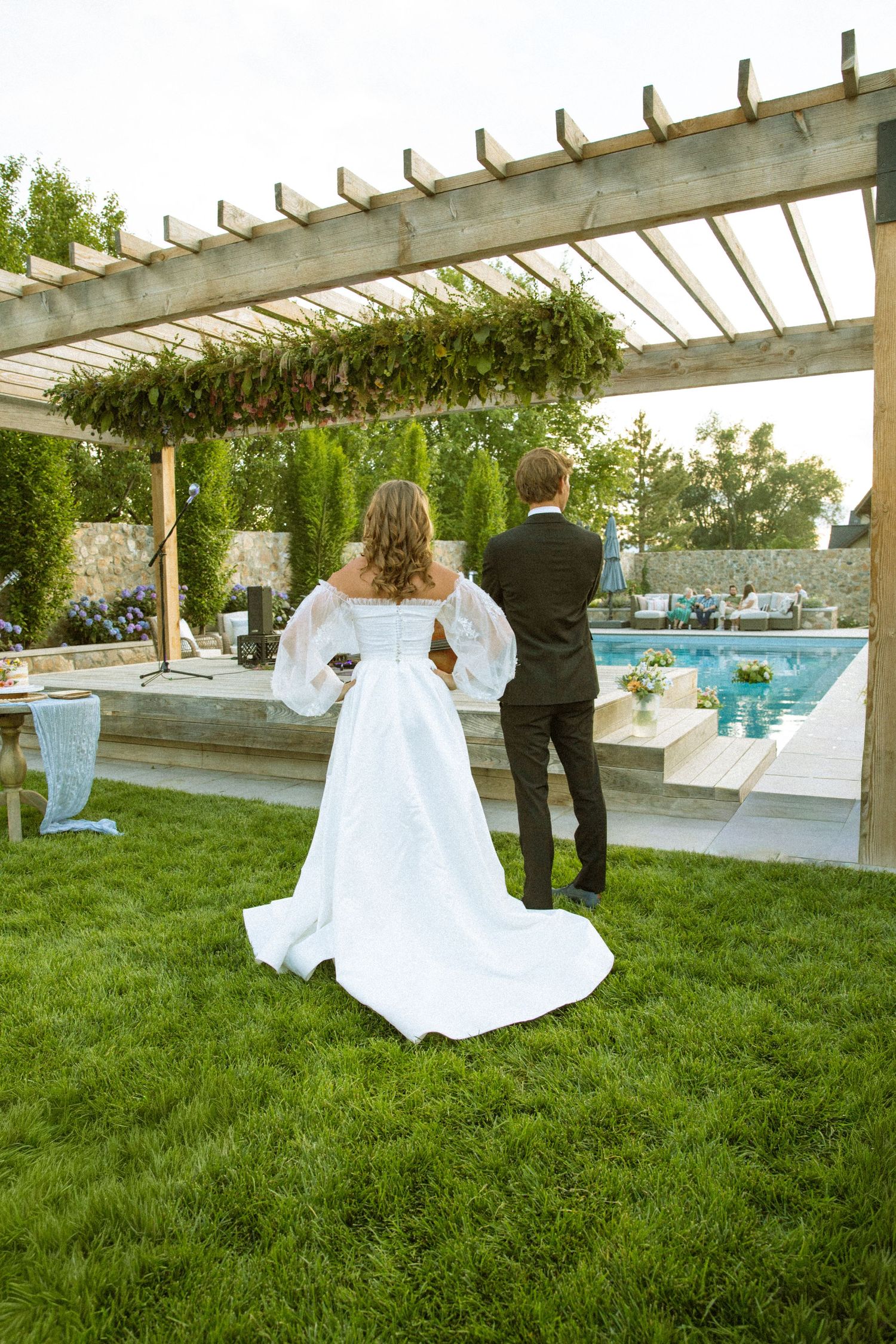 A bride in white dress walks across green lawn under a rustic wooden pergola.