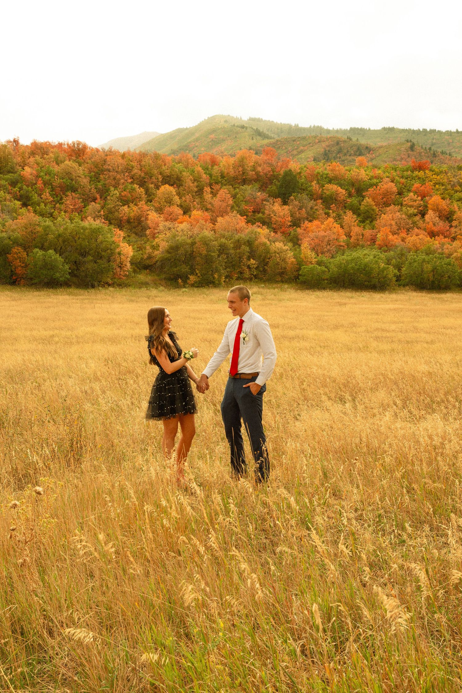 A couple stands together in a golden autumn field with vibrant fall foliage on rolling hills in the background.