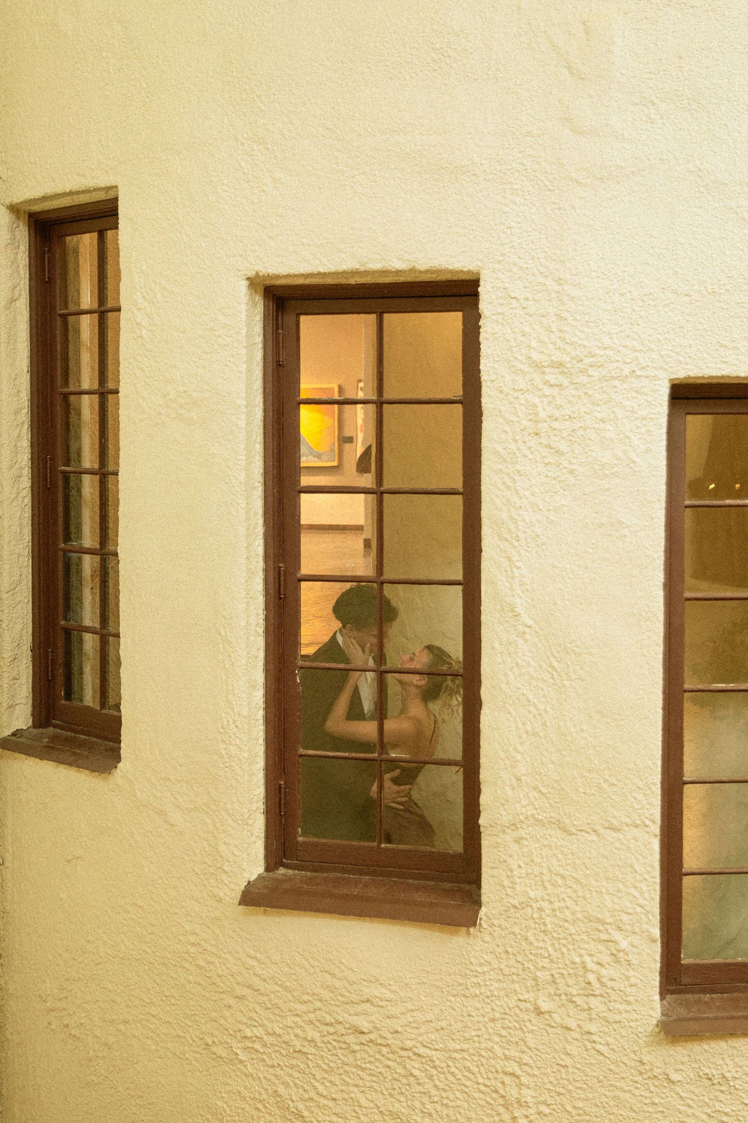 Through a window pane at night, a warm glow illuminates a figure seated at a table in a textured white building.