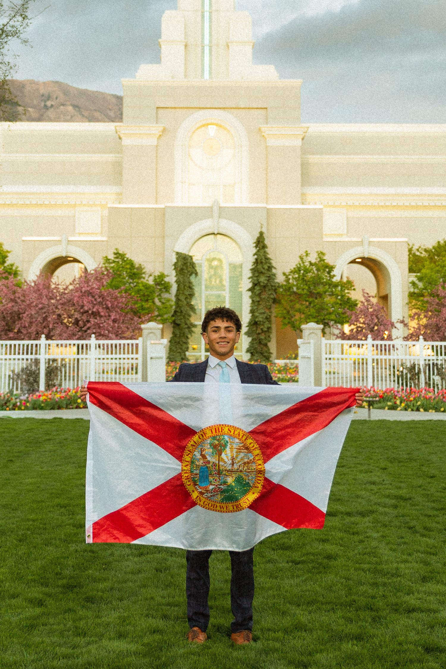 Young man in suit holding Florida state flag outside a white LDS temple with mountains in background.