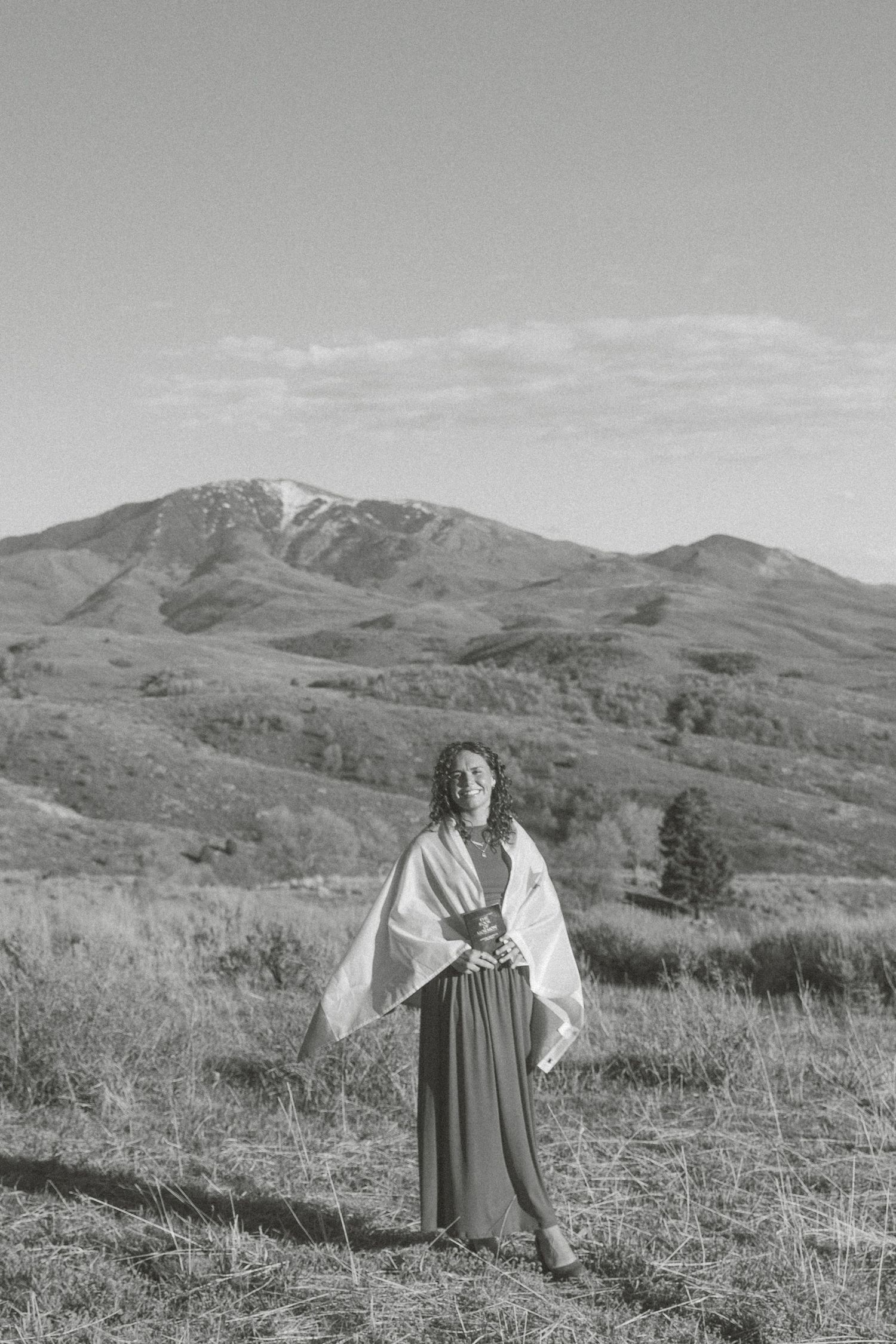 Black and white photo of a woman draped in a white shawl standing in a mountain meadow landscape.