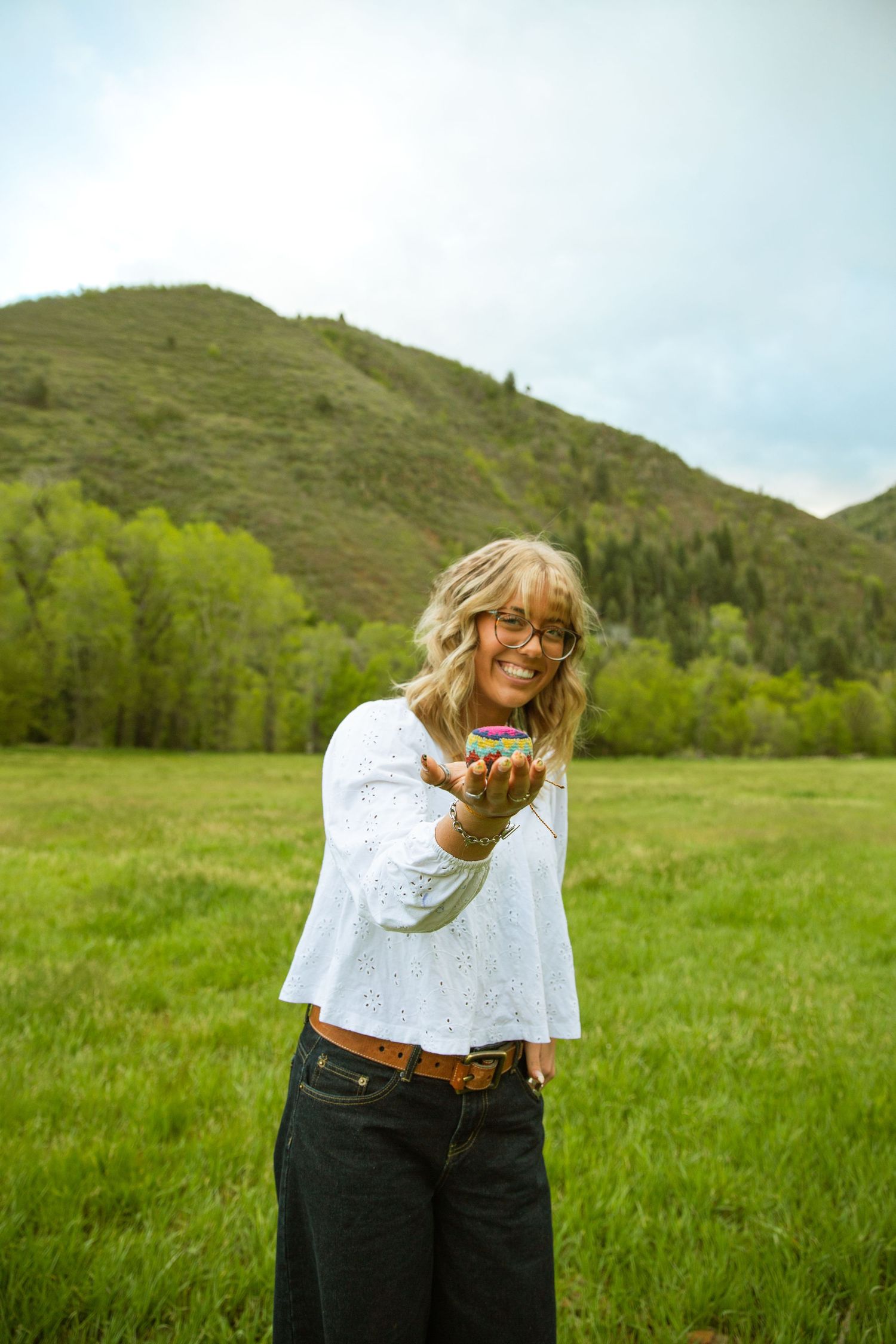 Smiling woman in white top holding small object outdoors in green mountain meadow landscape.