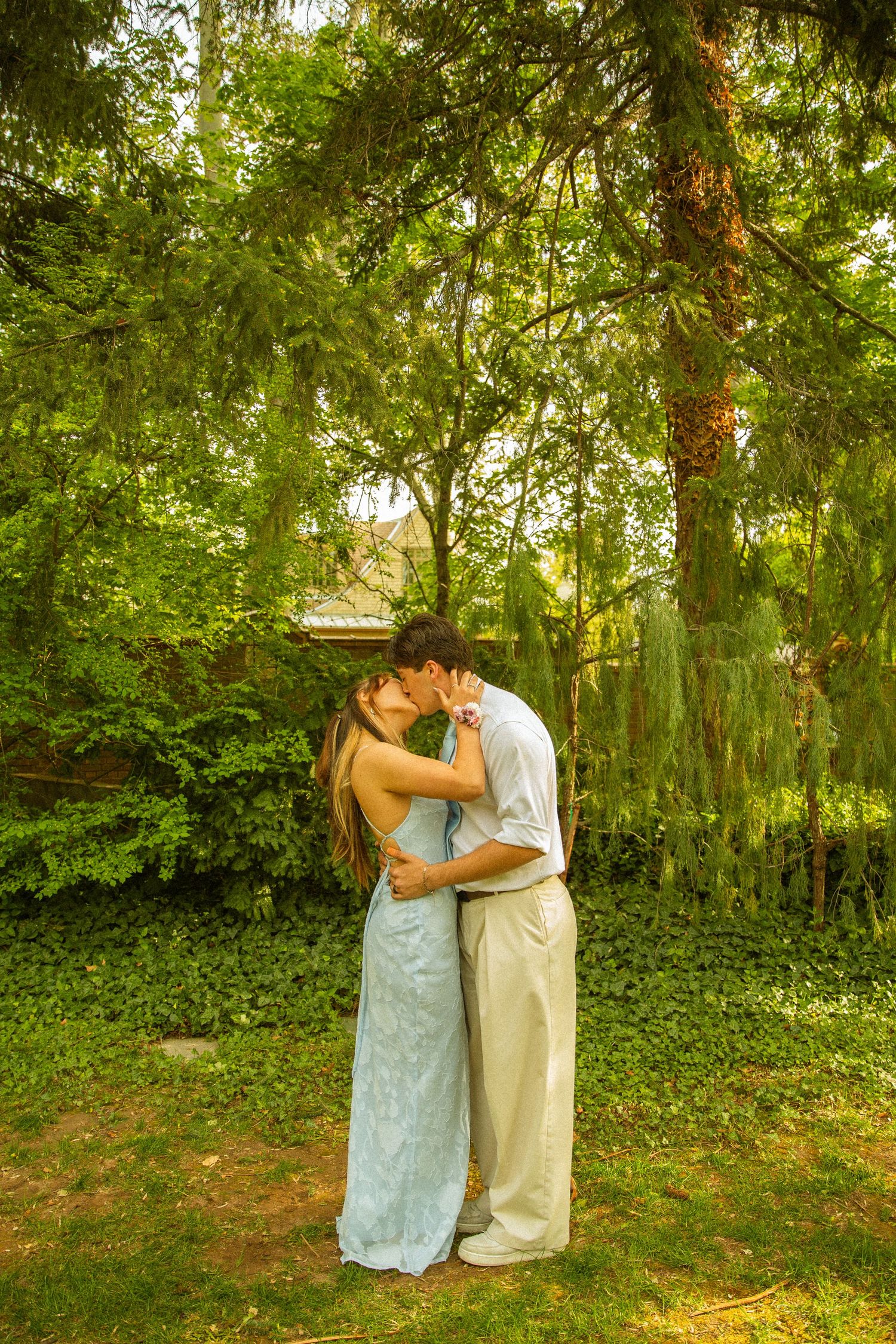A couple sharing a romantic kiss outdoors, surrounded by lush green trees, the woman in a light blue dress.