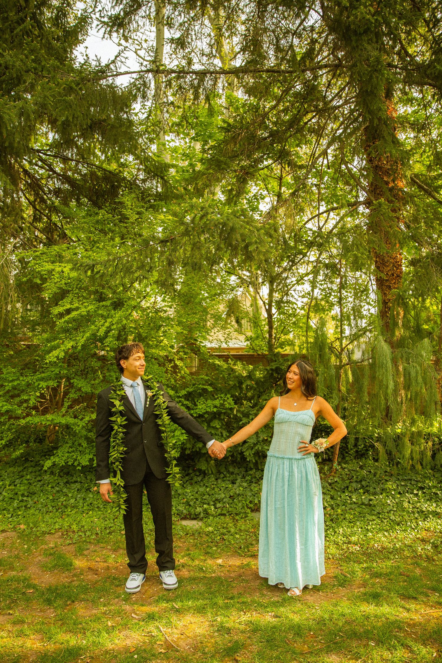 A young couple in formal prom attire holds hands outdoors, the boy in a black suit and girl in a light blue gown.