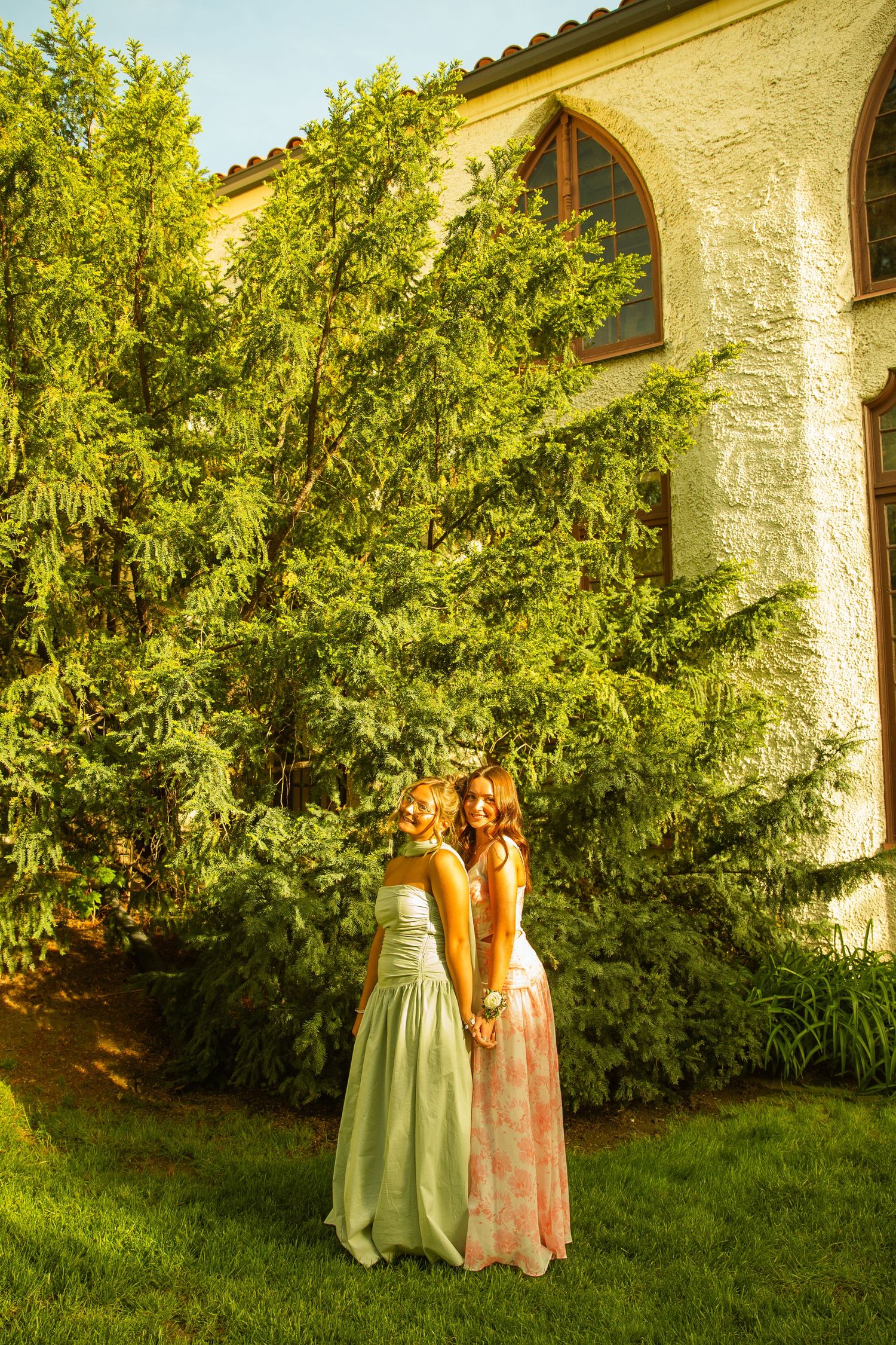 Two young women in elegant pastel gowns hold hands in a lush garden beside a charming stone building.