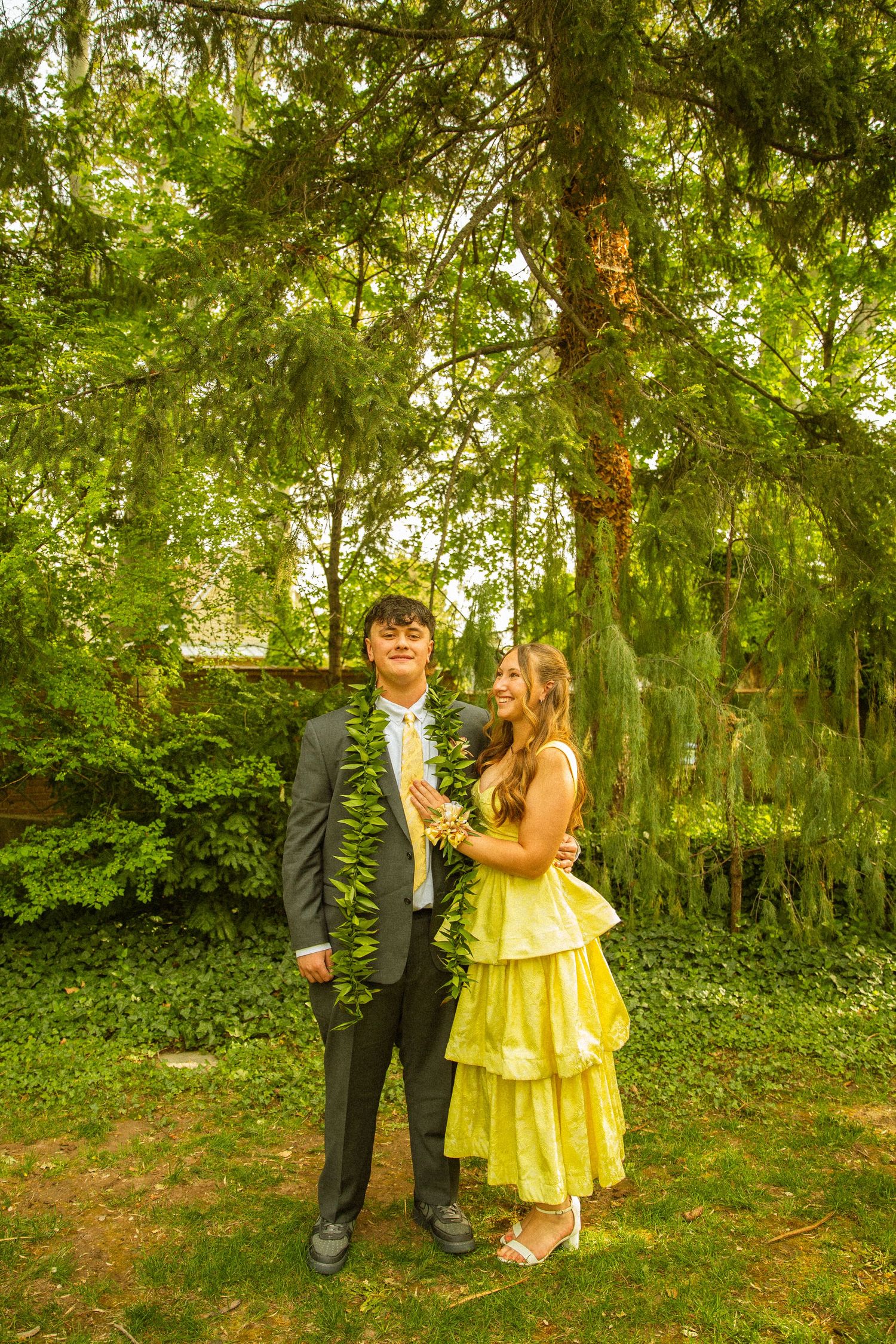 A couple in formal attire poses outdoors, the young man in a gray suit and the woman in a yellow tiered dress.