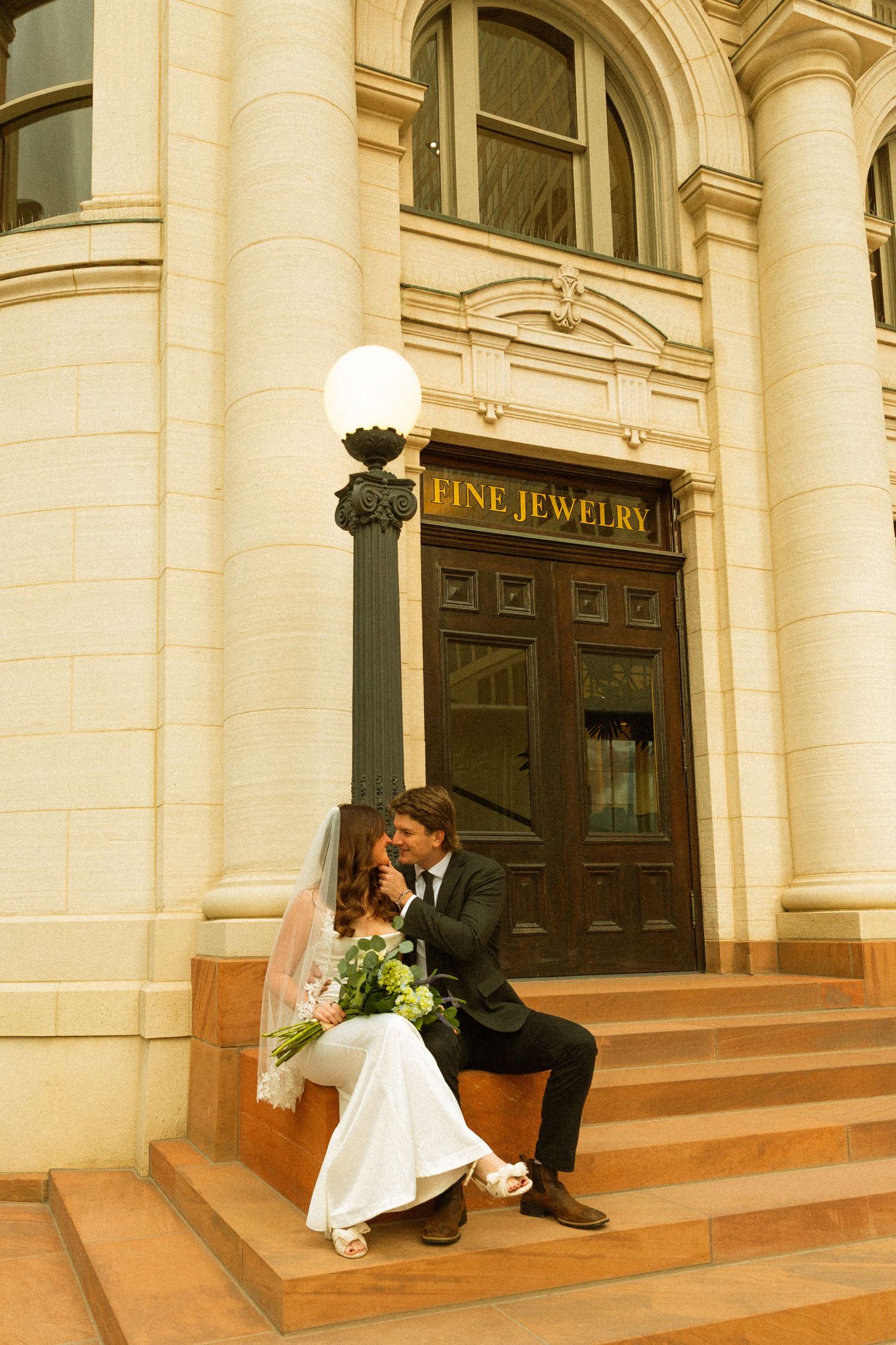 A couple in wedding attire sits on stone steps outside an ornate building with 