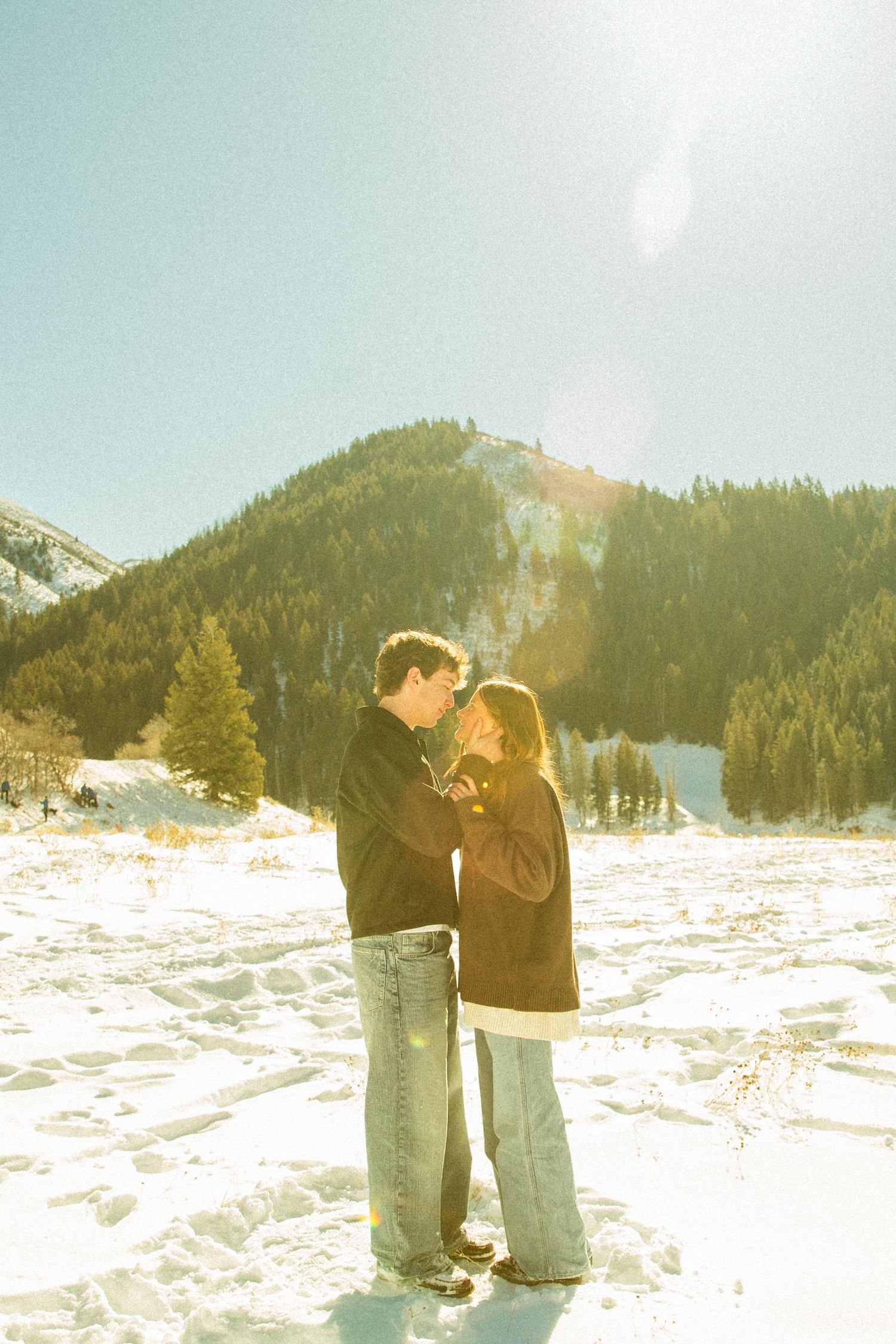 A couple embraces in a snowy mountain landscape on a sunny winter day with evergreen trees in the background.