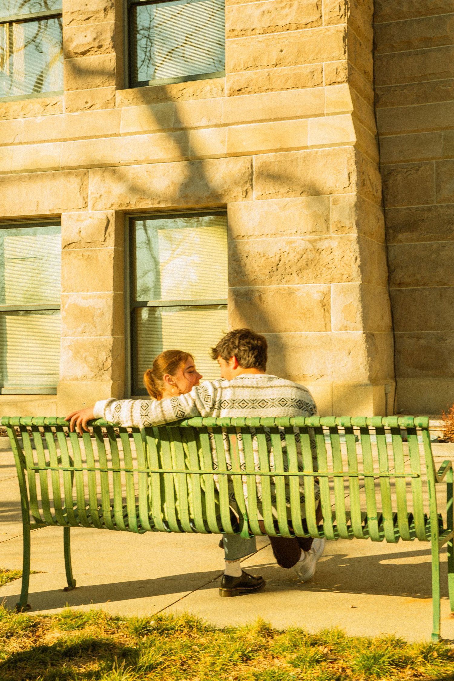 Two people sit together on a green bench outside a stone building on a sunny day.