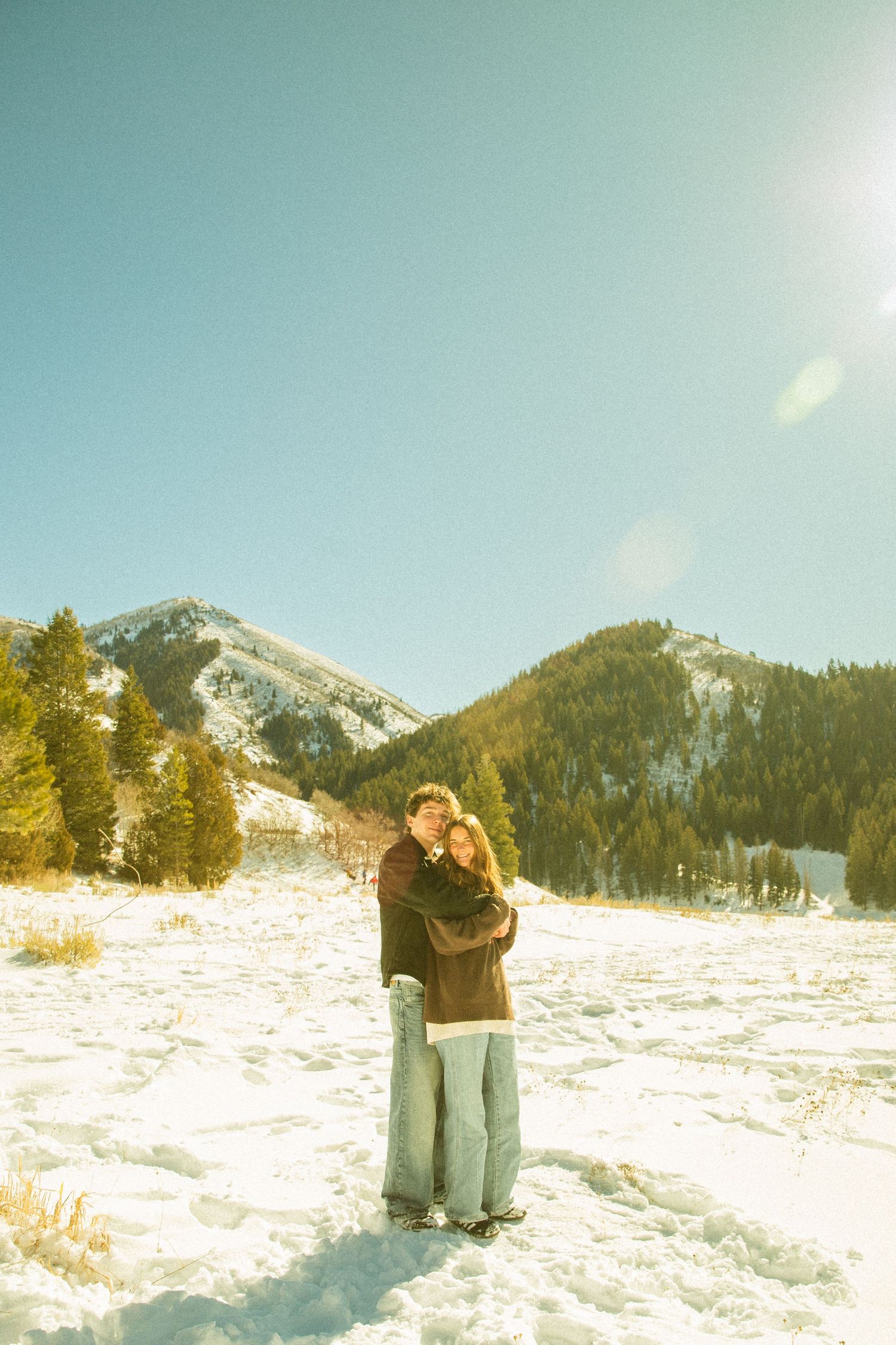 A couple embraces in a snowy mountain landscape with evergreen trees and sunlit peaks in the background.