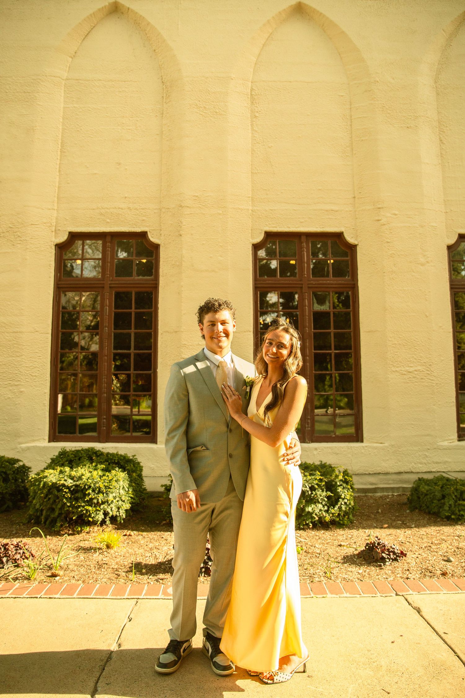 Couple in formal attire posing outside a white brick building, man in gray suit, woman in gold gown.