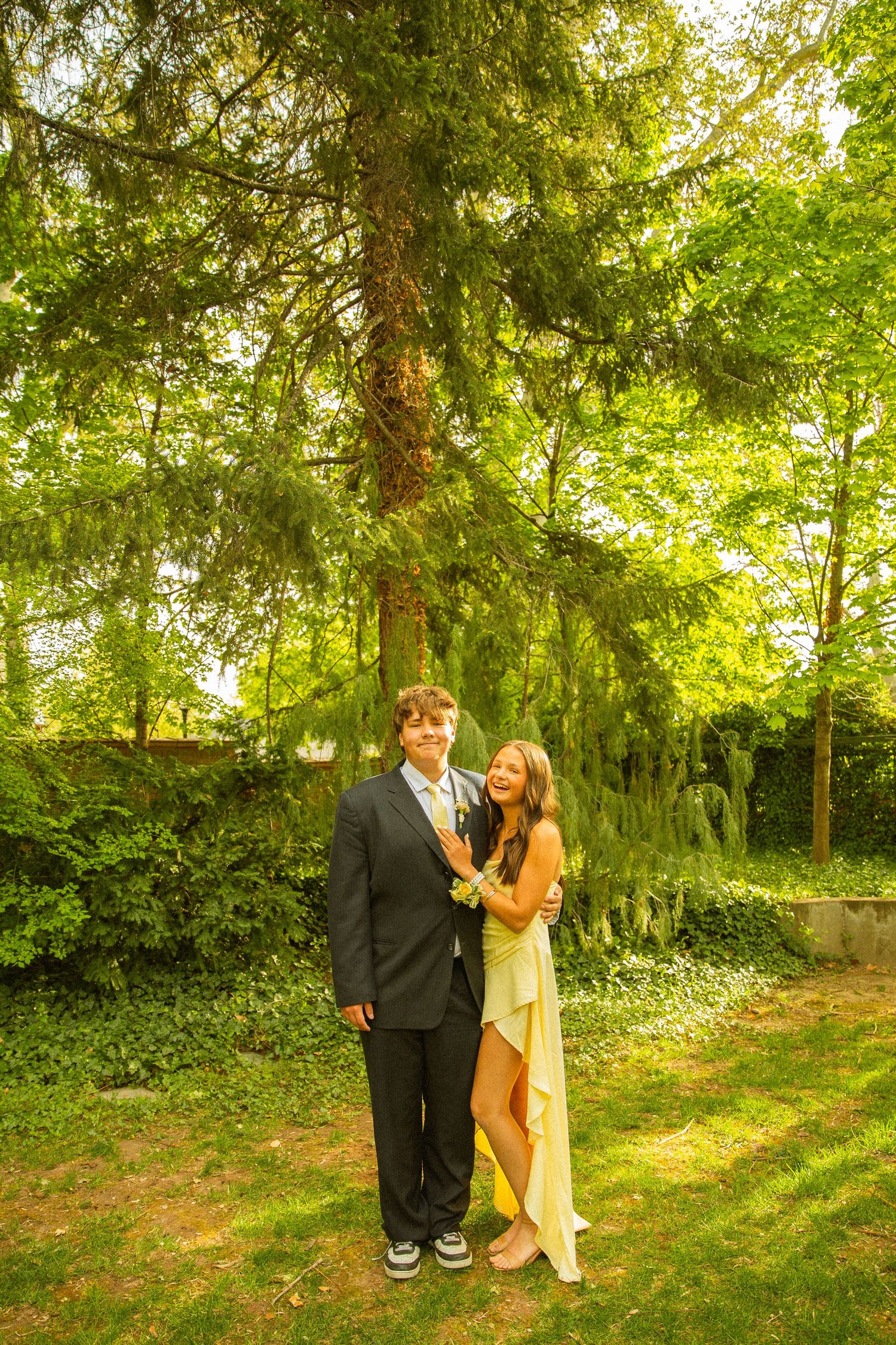A young couple dressed in prom attire pose outdoors, the girl in a gold dress and the boy in a dark suit.