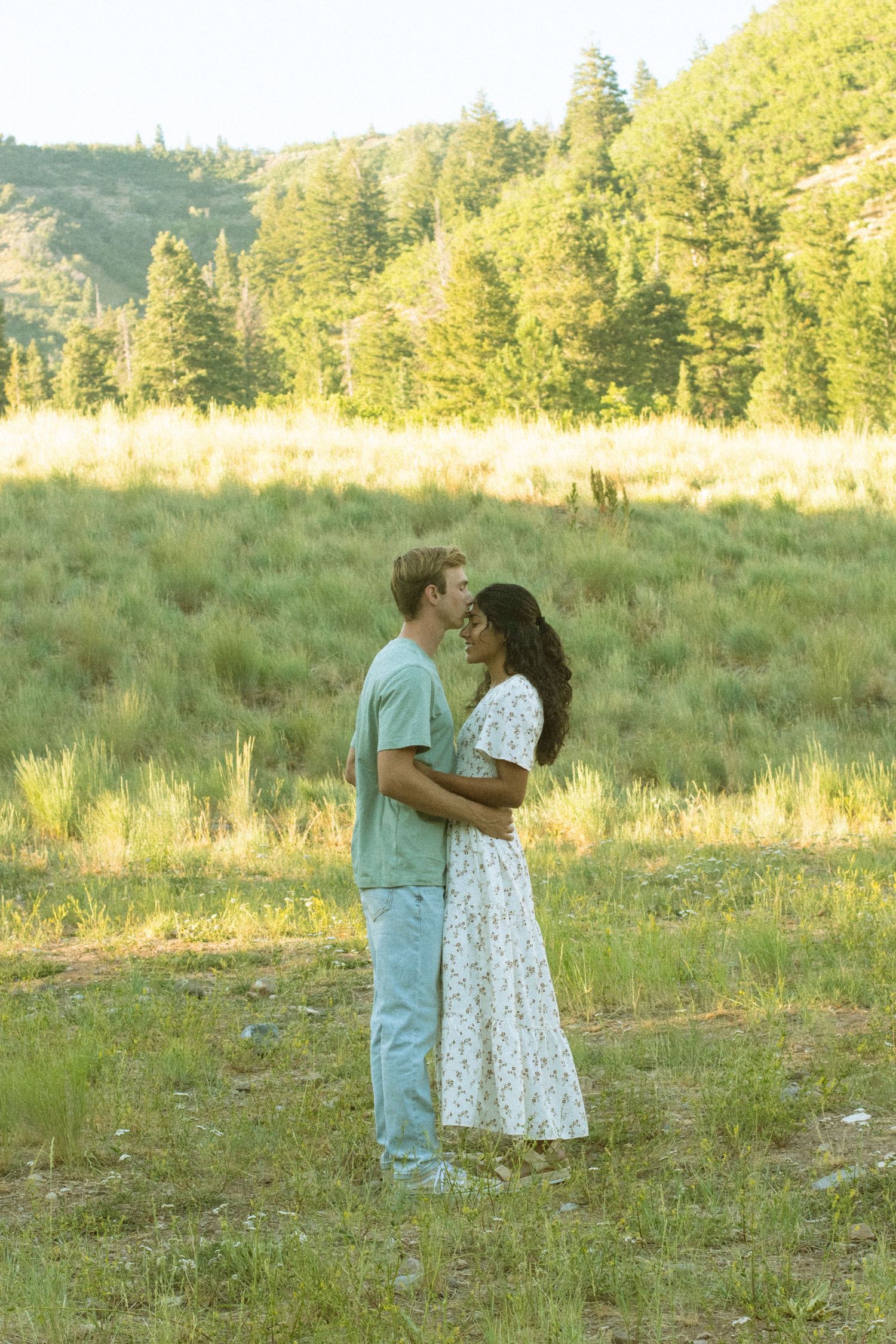 A couple shares a tender moment in a sunlit meadow surrounded by evergreen trees during golden hour.
