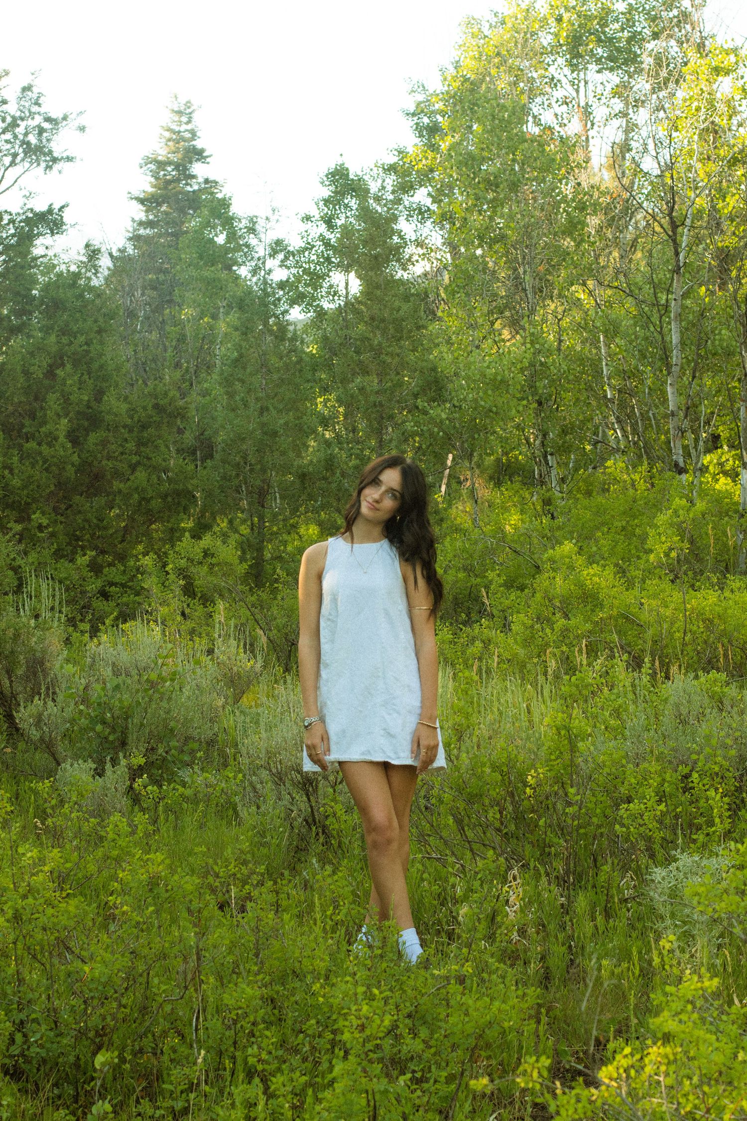 Person in a light blue sleeveless top poses among lush green foliage and wildflowers in a natural outdoor setting.