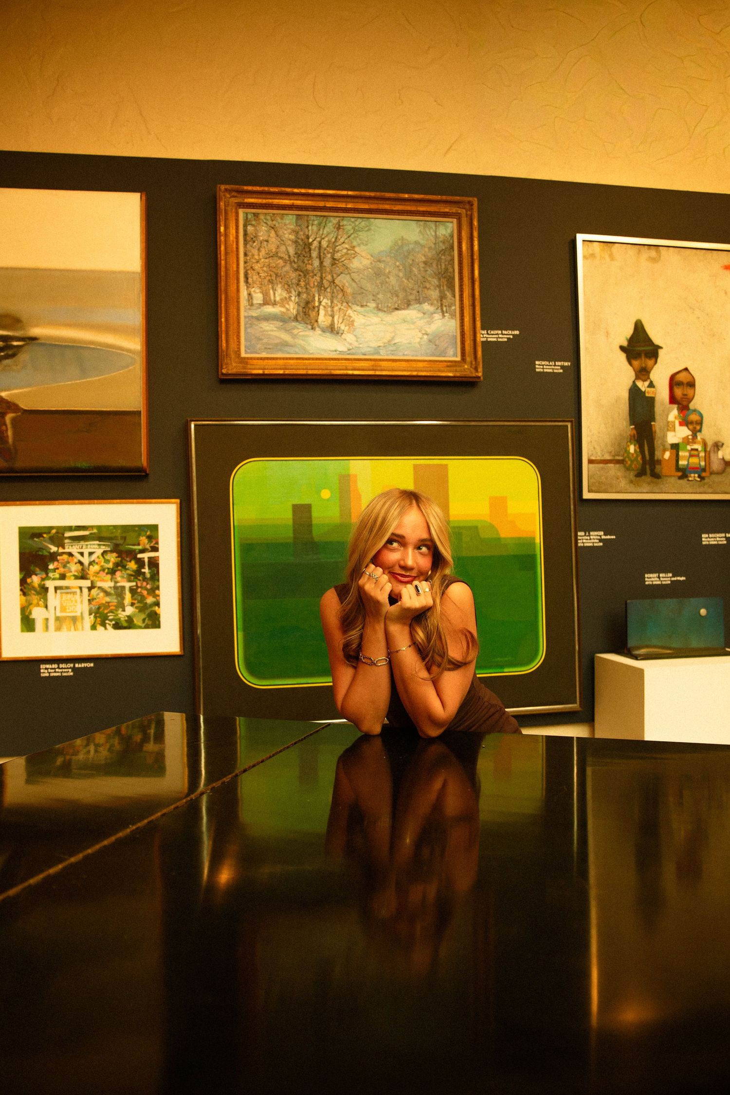 A smiling blonde woman poses on a reflective floor surrounded by colorful artwork in a dimly lit art gallery.