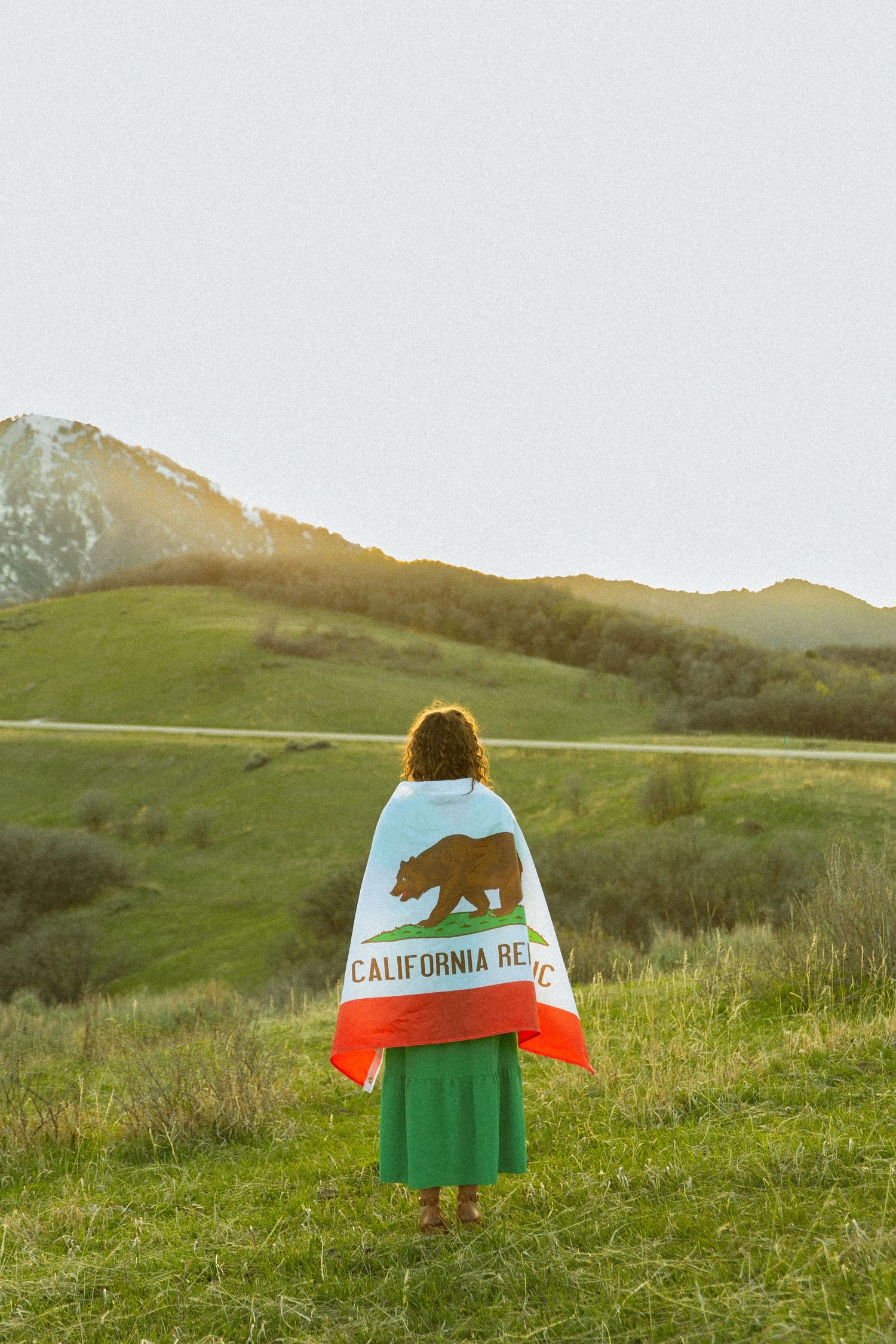 Person wrapped in California Republic bear flag standing in green meadow with scenic mountain backdrop at sunset.