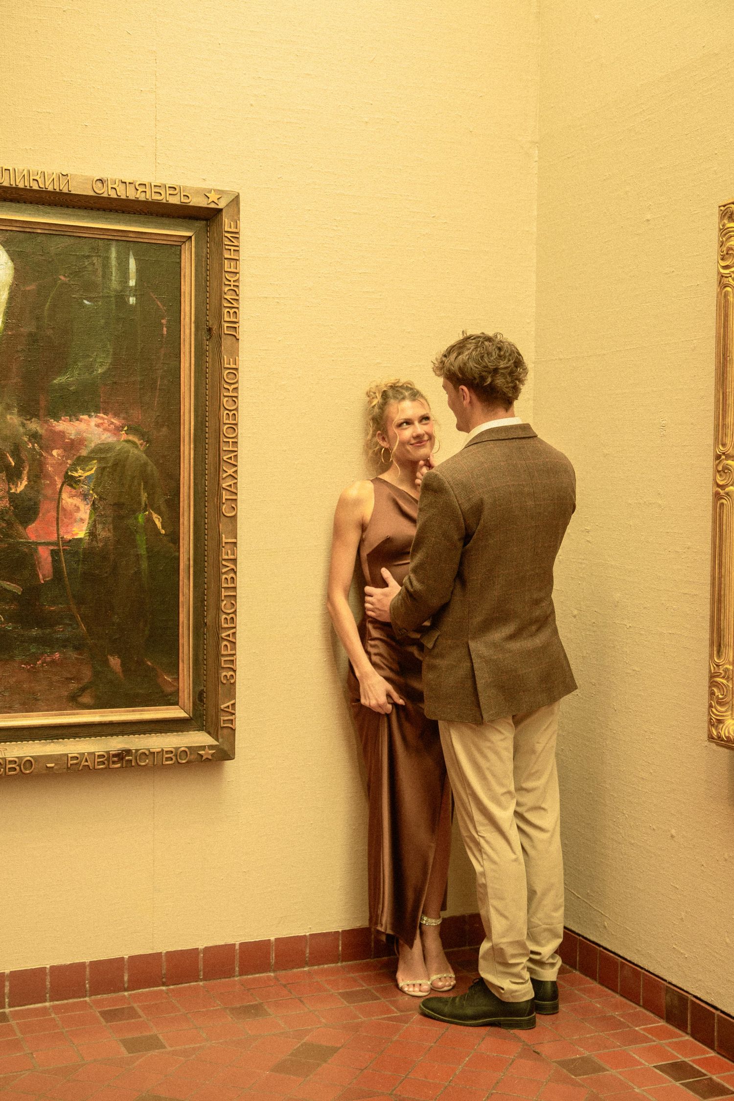 Two people stand close together in an art gallery with gold-framed paintings on cream-colored walls and terracotta flooring.
