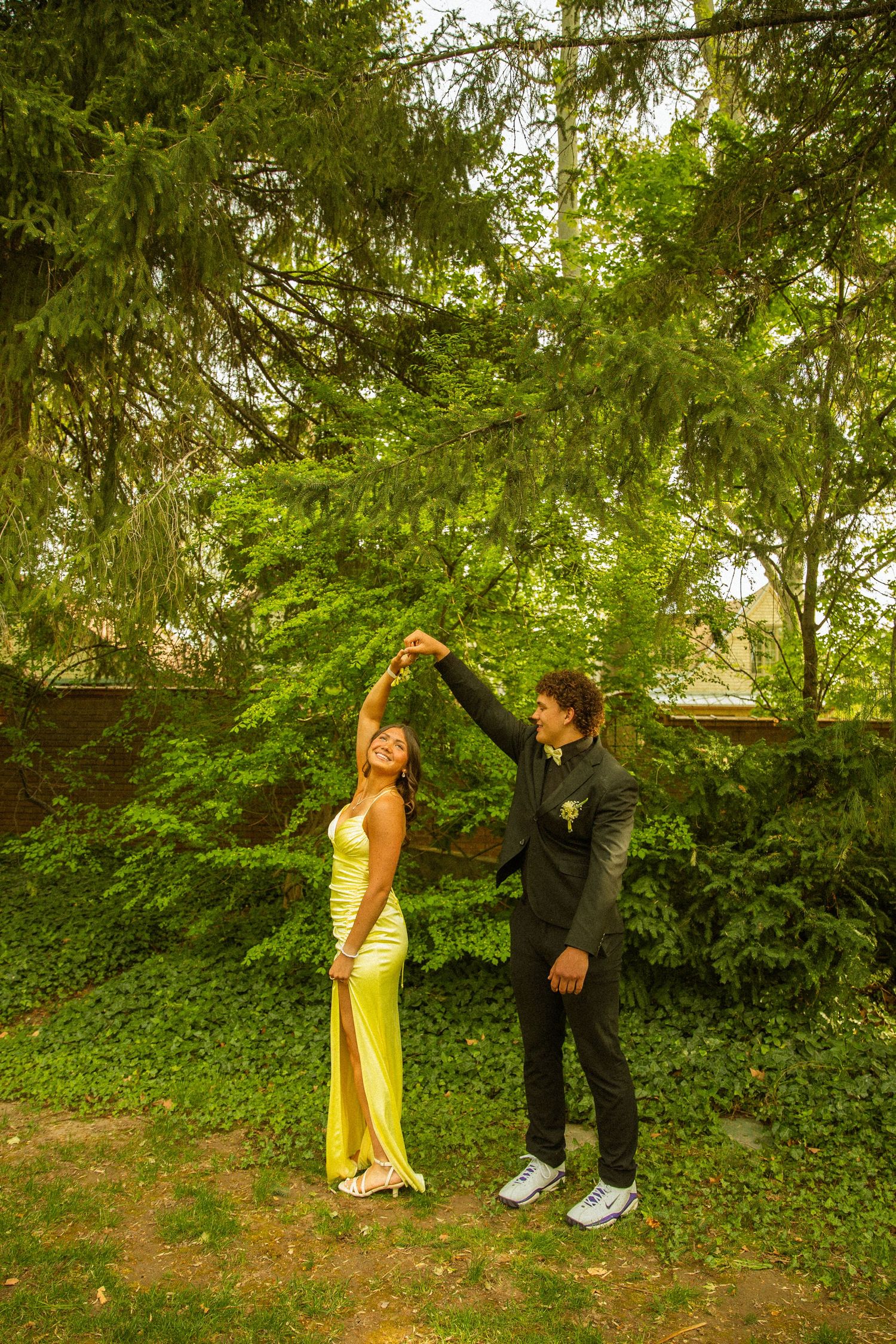 A couple in formal prom attire dancing outdoors, the woman in a yellow gown being twirled by her partner in a black suit.
