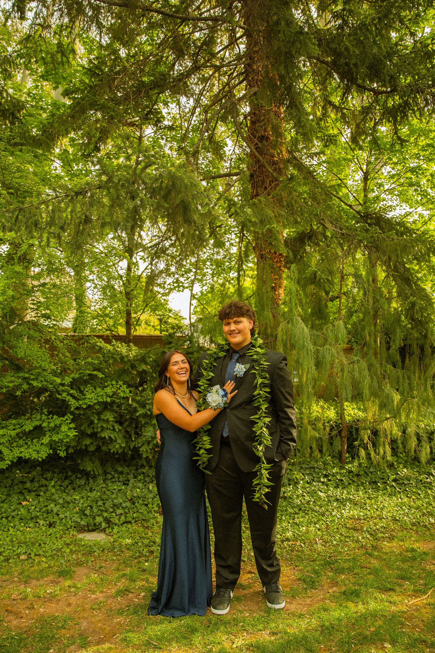A couple in formal prom attire poses outdoors under lush green trees, wearing corsage and boutonniere.