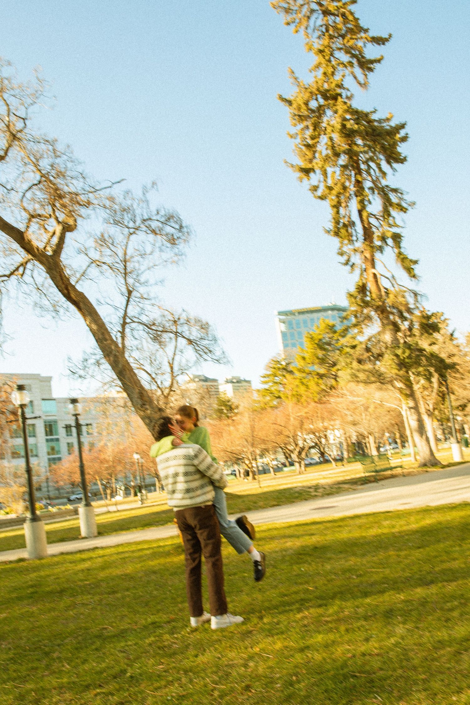A couple embraces in a warm sunset glow on a grassy lawn with evergreen trees and buildings in the background.