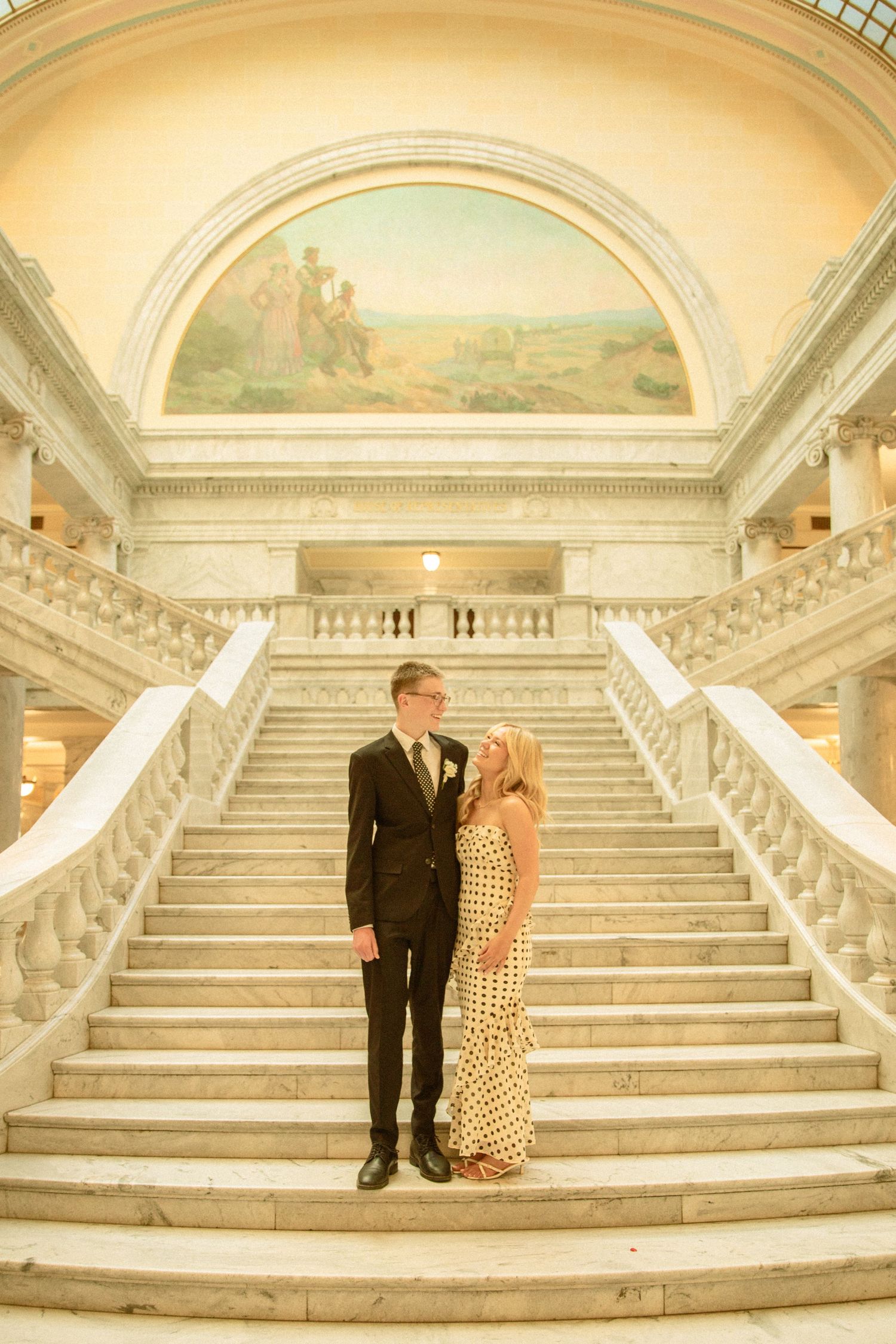 Elegant couple stands on grand marble staircase in historic building with arched mural ceiling and ornate architecture.