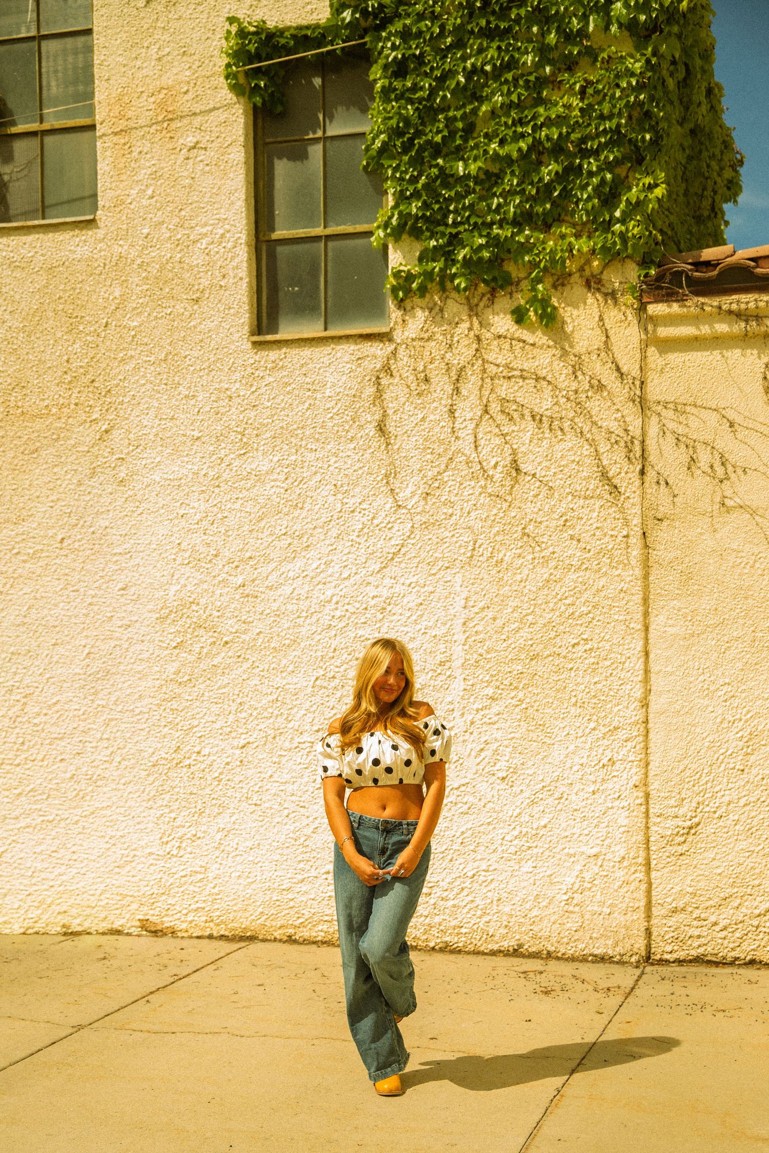 Woman in polka dot crop top and wide-leg jeans posing against a textured white wall with ivy.
