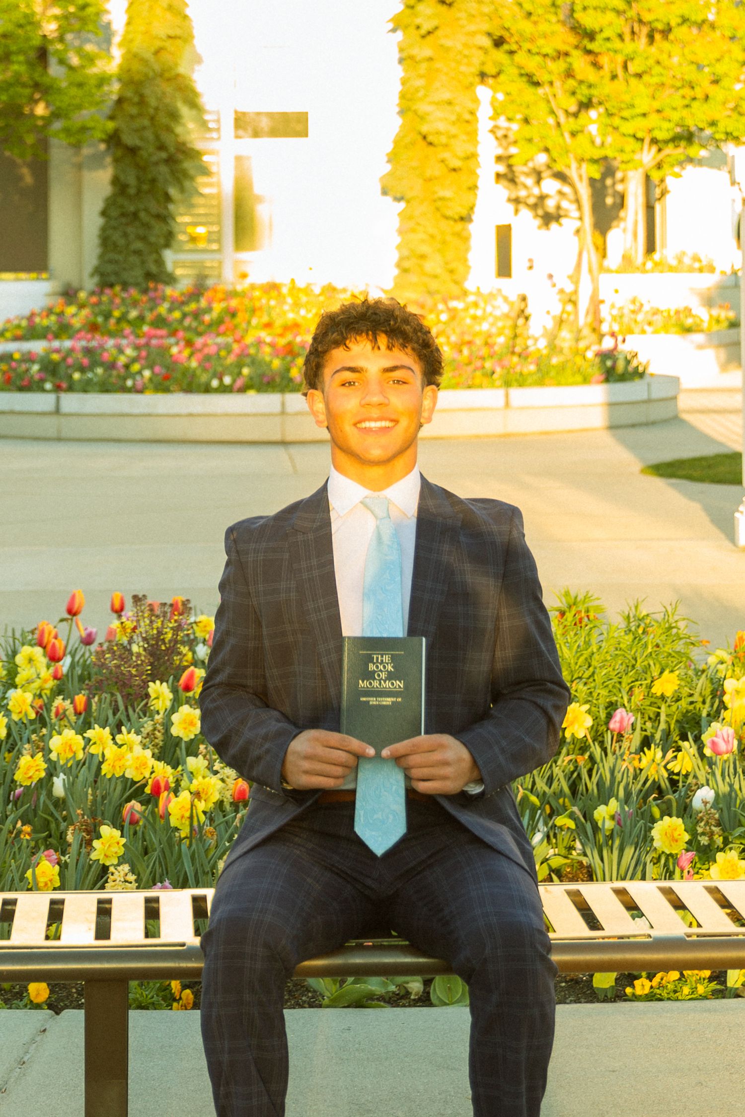 Young man in plaid suit holding Book of Mormon sits on bench surrounded by colorful tulips and daffodils.