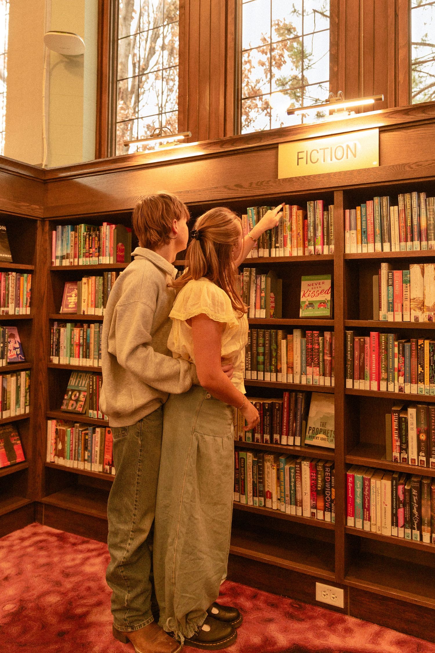 Students browse books on wooden library shelves with red carpeting in a warm-lit academic setting.