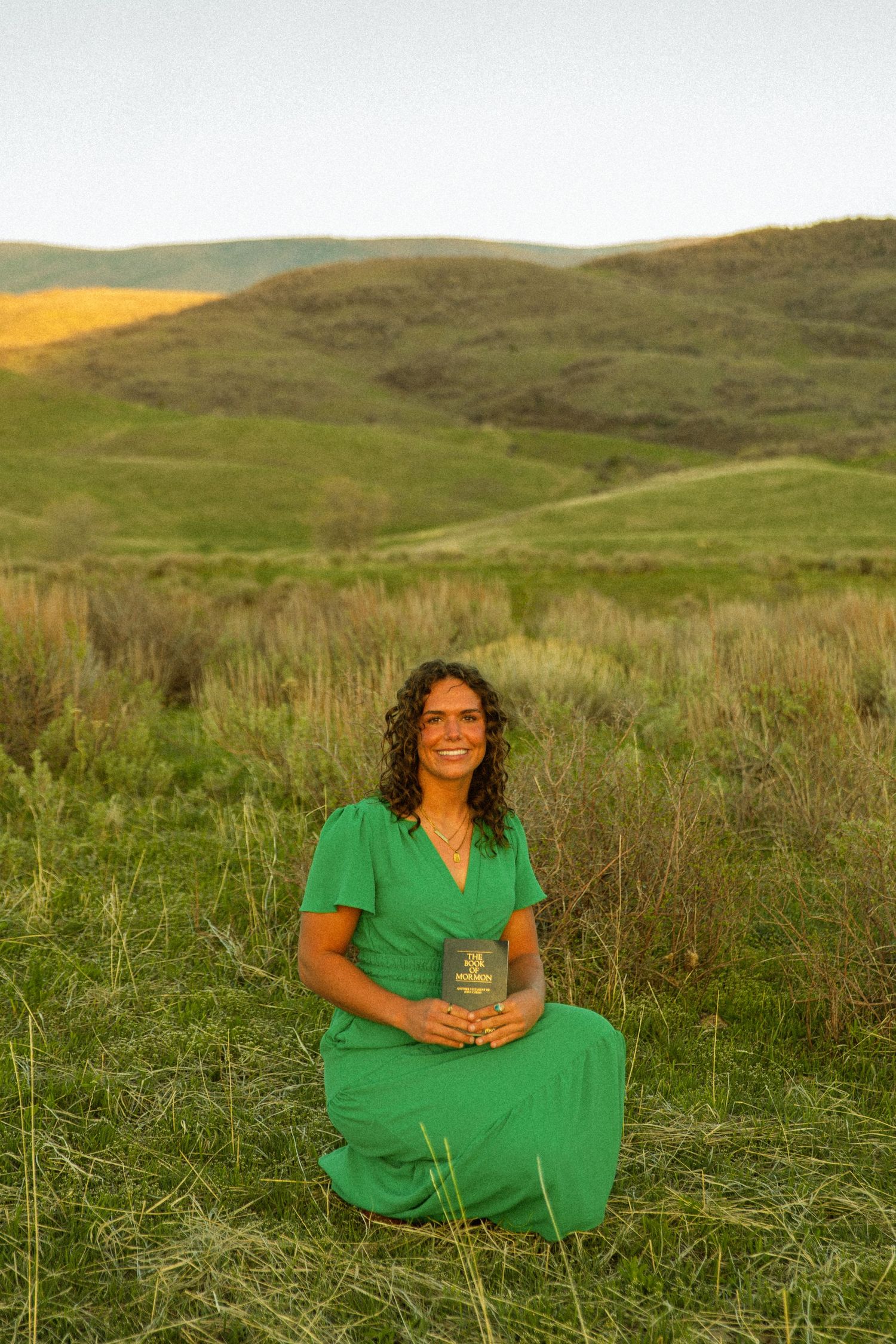Woman in green dress sitting in lush meadow holding a book, with rolling green hills in golden sunset light.