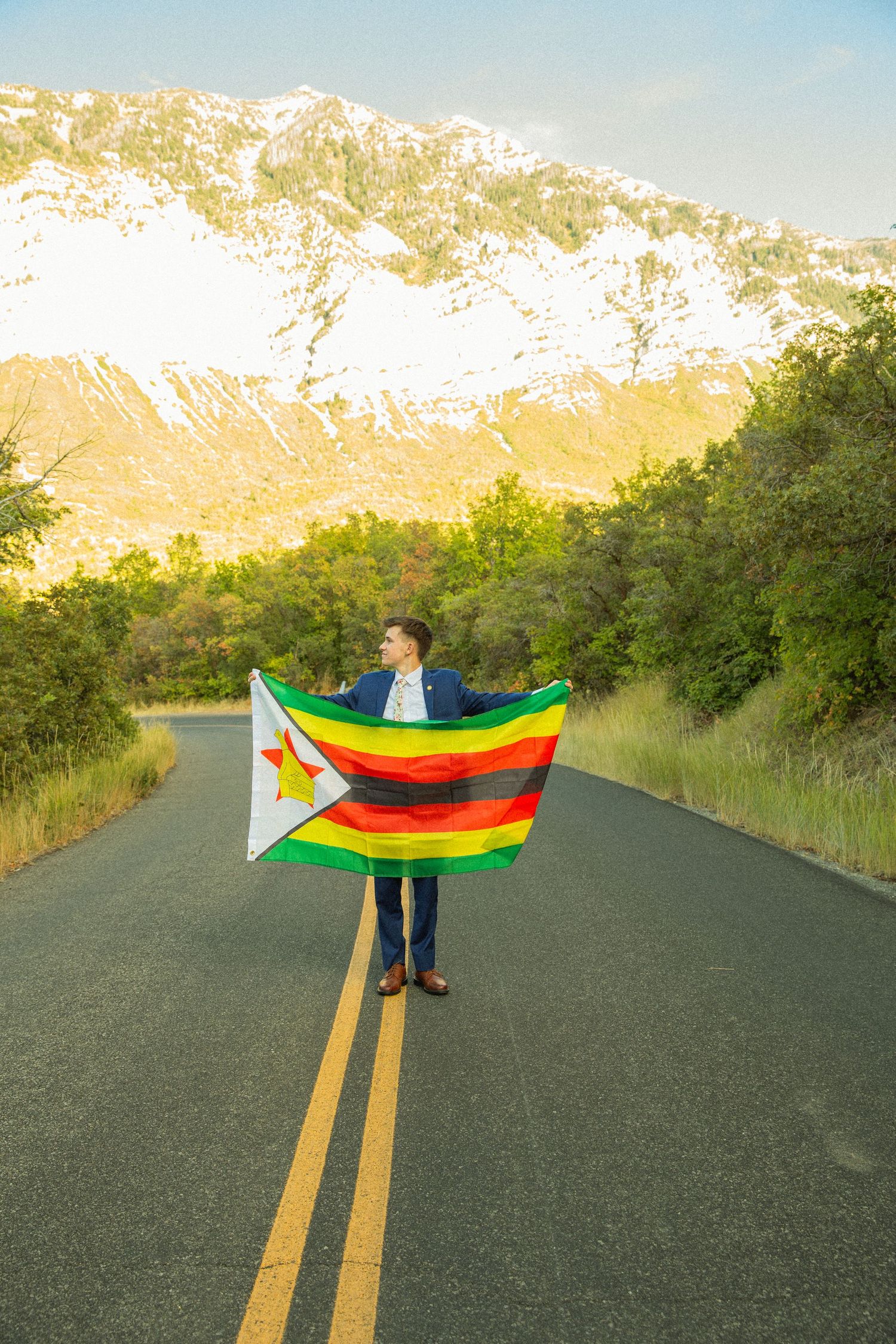 Person standing in middle of empty road holding colorful flags against mountain backdrop at sunset.