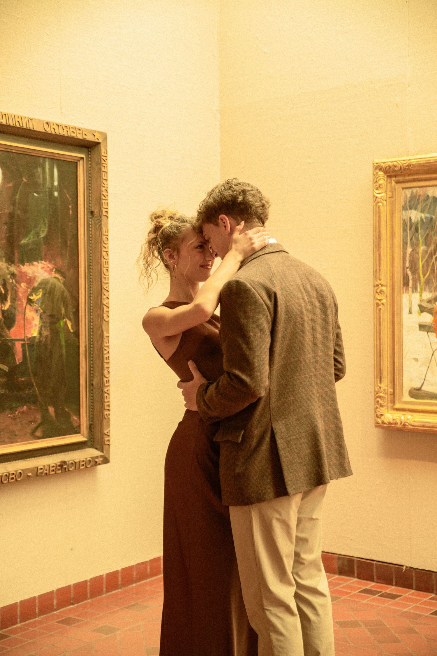 A couple views artwork in a gallery, walking along a wall displaying framed paintings in warm lighting.