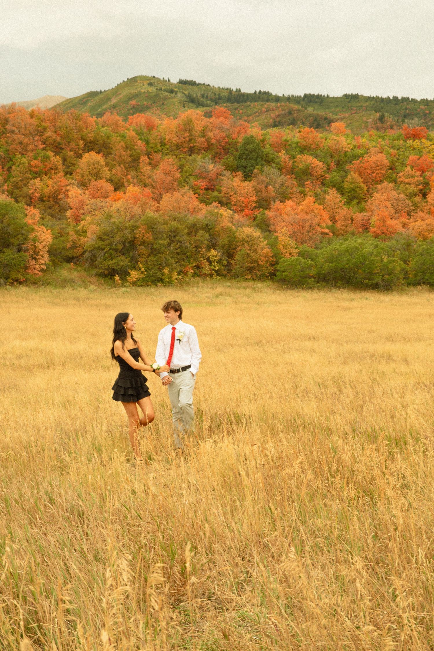 A couple stands together in a golden autumn meadow with vibrant orange foliage on rolling hills in the background.