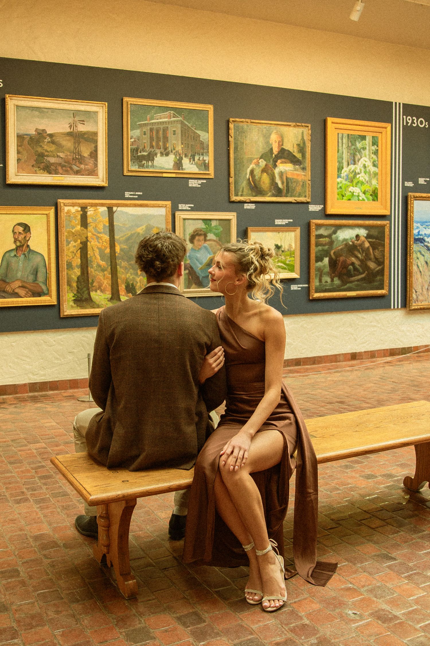 Two people sit together on a wooden bench viewing artwork displayed in a gallery with warm lighting and brick floors.