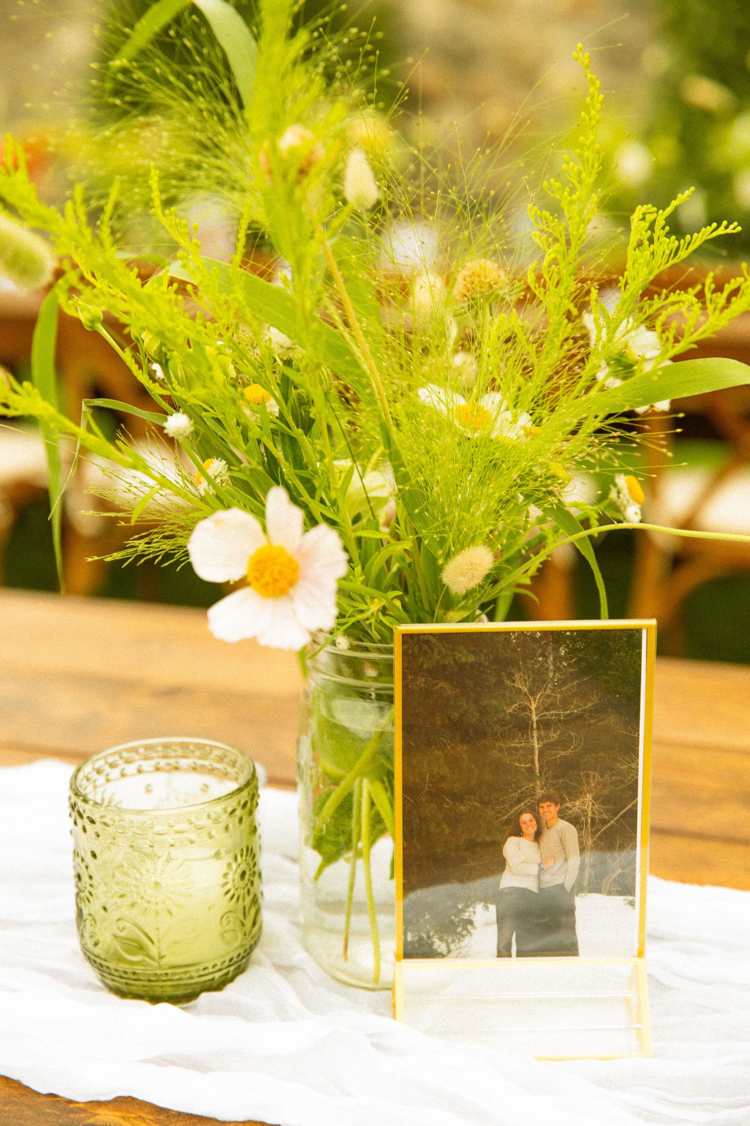 Elegant floral centerpieces with white flowers and greenery in green glass vases on a white tablecloth.