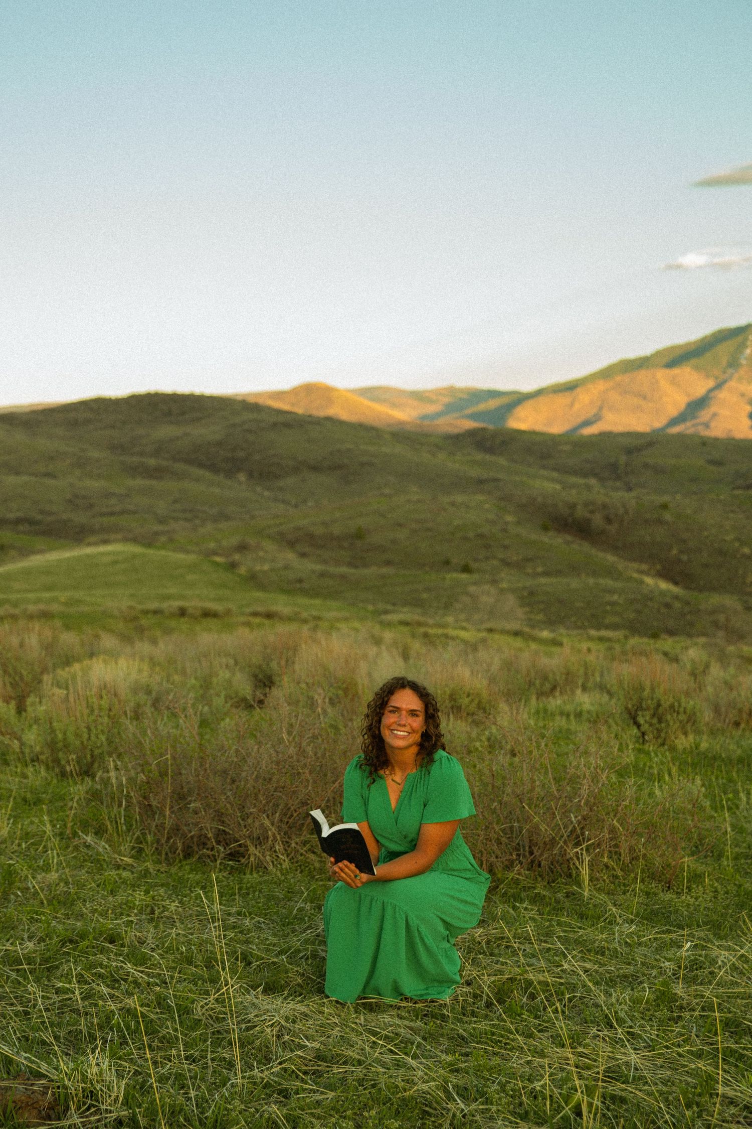 Woman in green dress reading a book while kneeling in a lush green meadow with rolling hills at sunset.