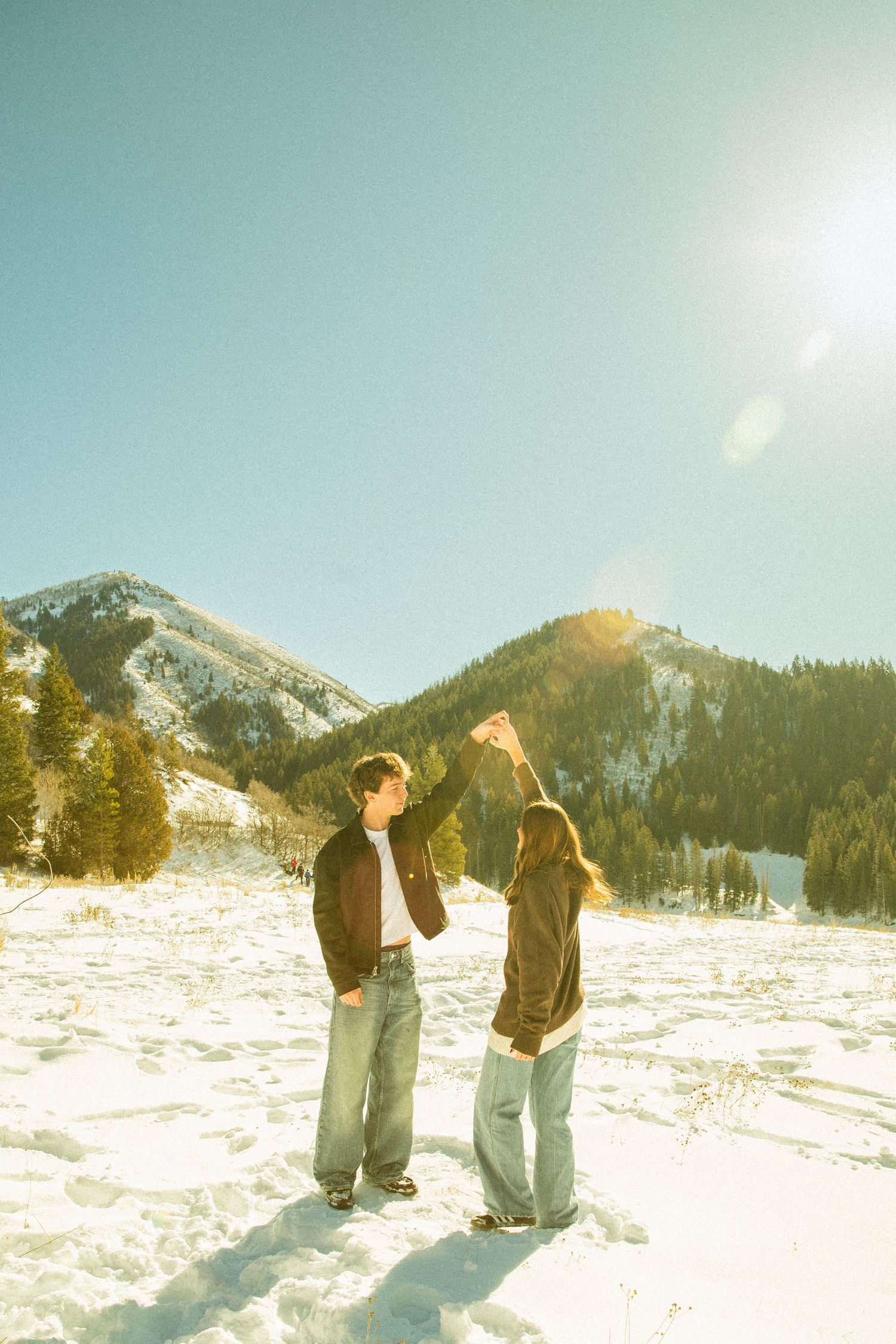 Two people playing in deep snow with snow-capped mountains and evergreen forest in the background.