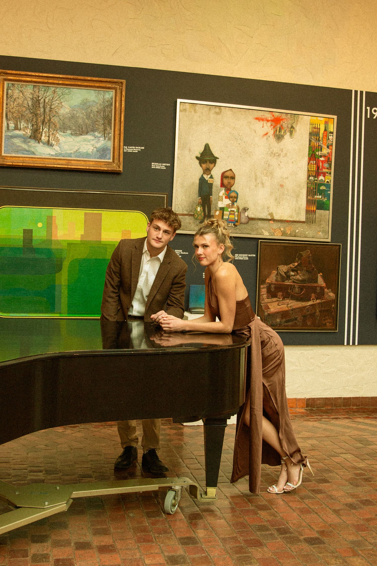 Two people pose elegantly by a grand piano in an art gallery with paintings displayed on dark walls behind them.