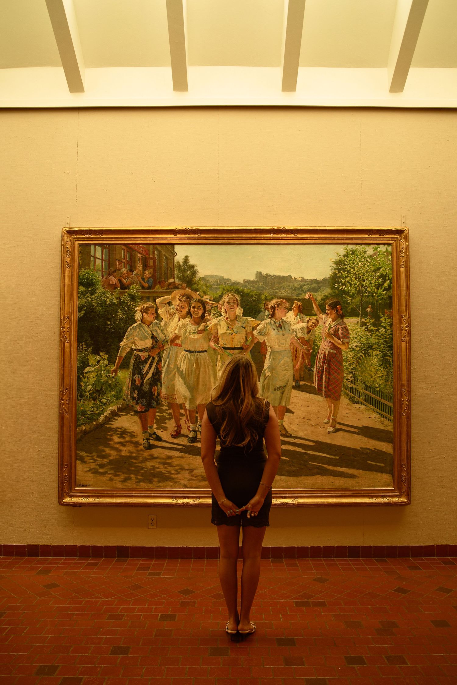 A woman stands before a large framed painting of women in white dresses in a warmly lit art gallery.