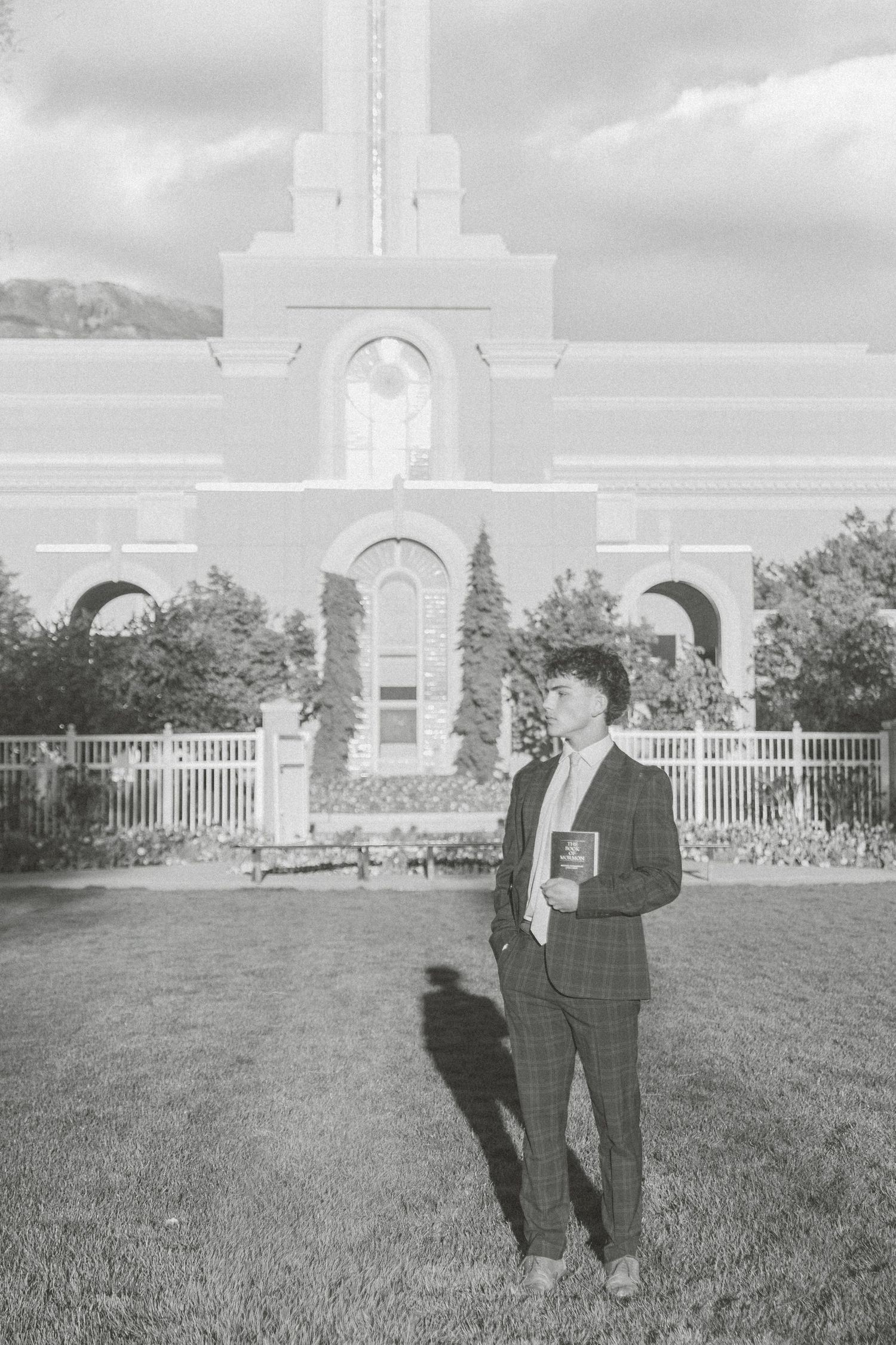 Young man in suit holding book stands on grass before a grand white church building in black and white photo.