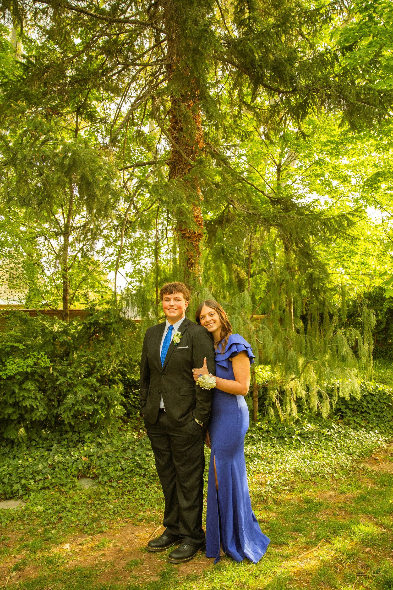 A young couple dressed in prom attire, a black suit and blue gown, pose together outdoors under lush green trees.