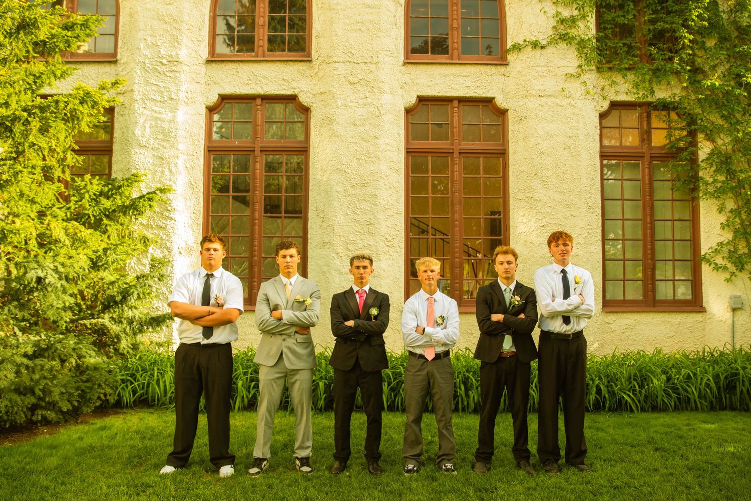 Six young men in formal attire stand with arms crossed in front of a brick building surrounded by greenery.
