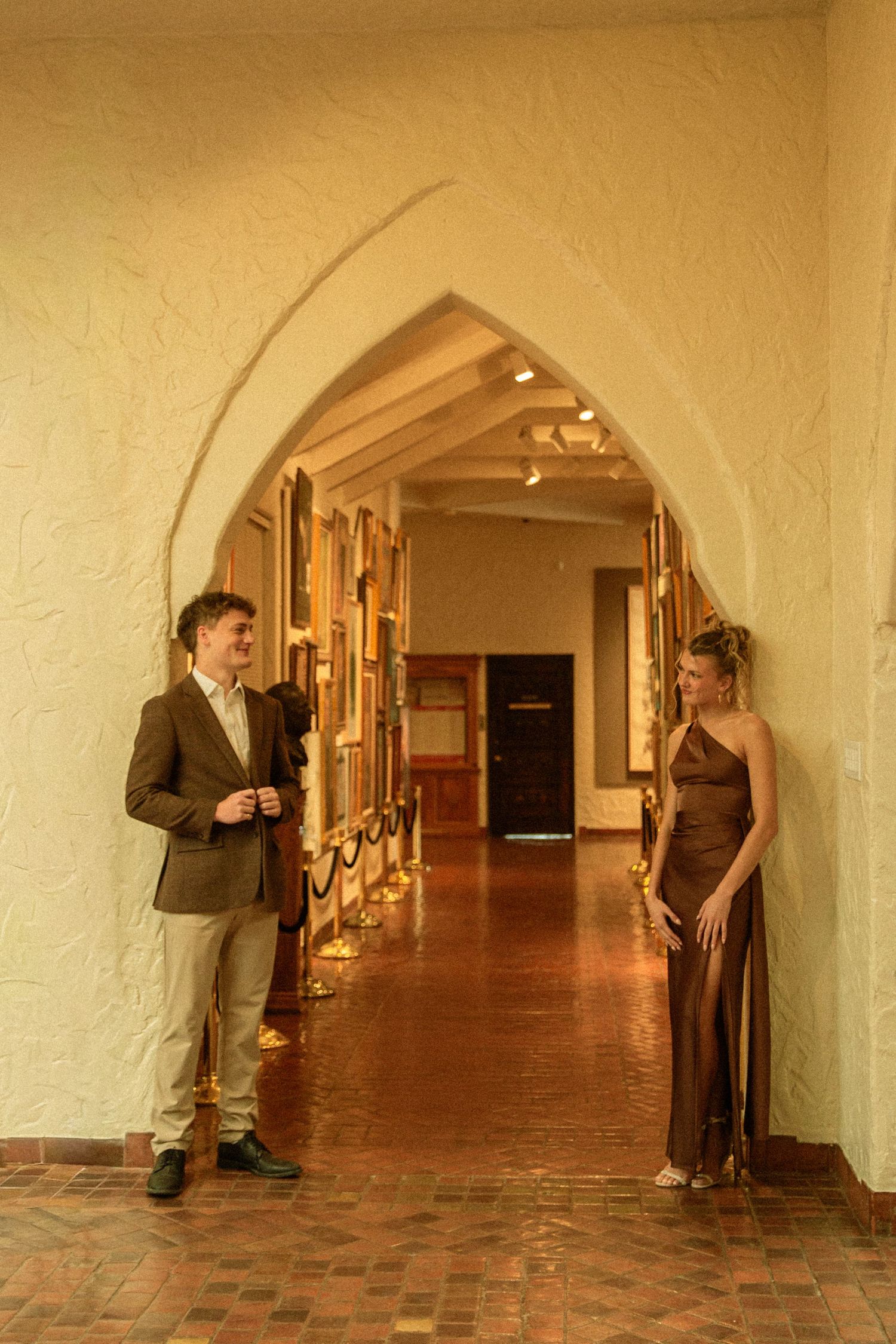 Two people stand in an elegant arched hallway with warm lighting and brick flooring at a formal event.
