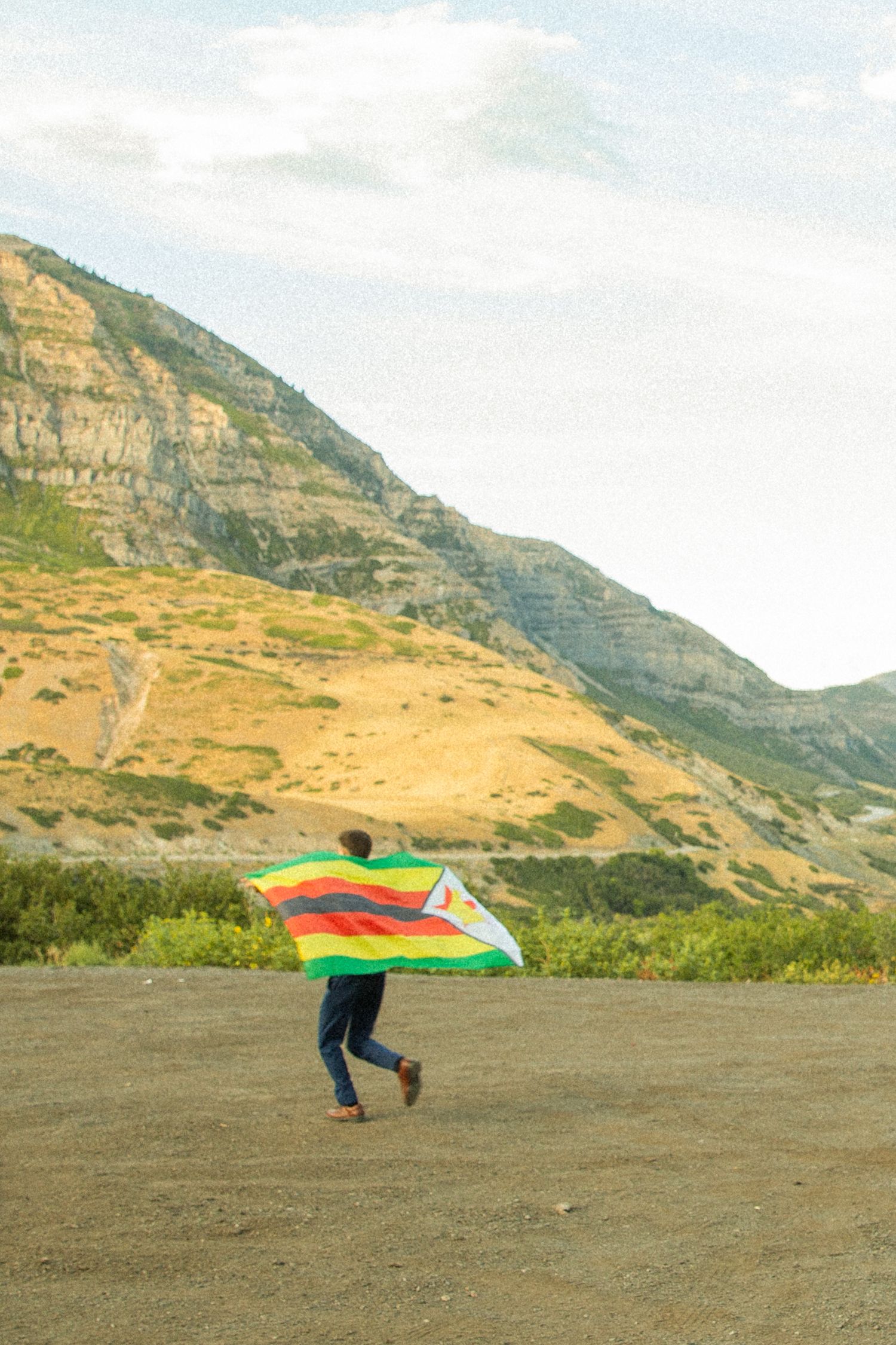 A person runs with a colorful kite against a backdrop of rugged mountain cliffs and golden hills.